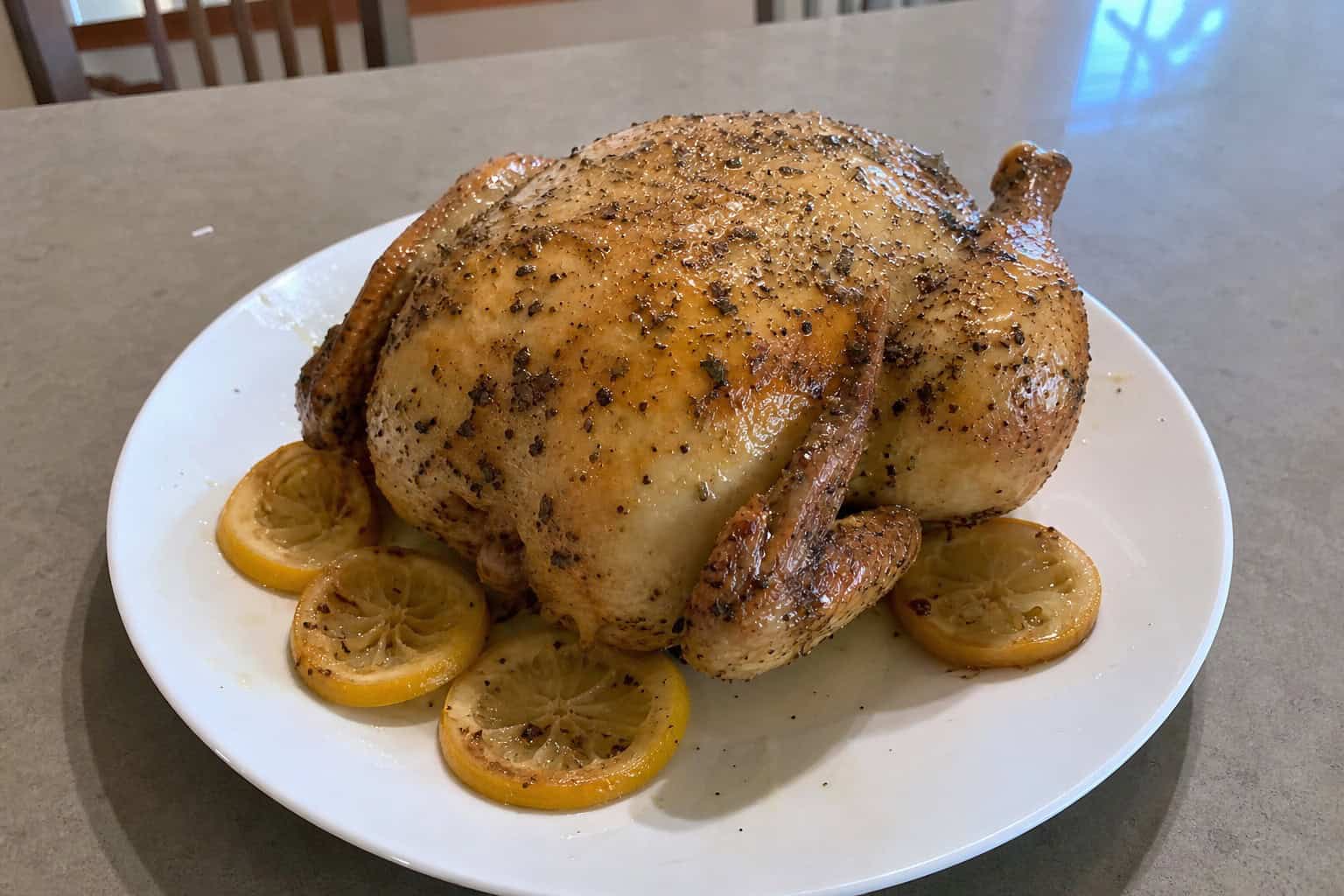 A whole roasted chicken garnished with lemon slices and fresh herbs on a wooden board in a kitchen setting.