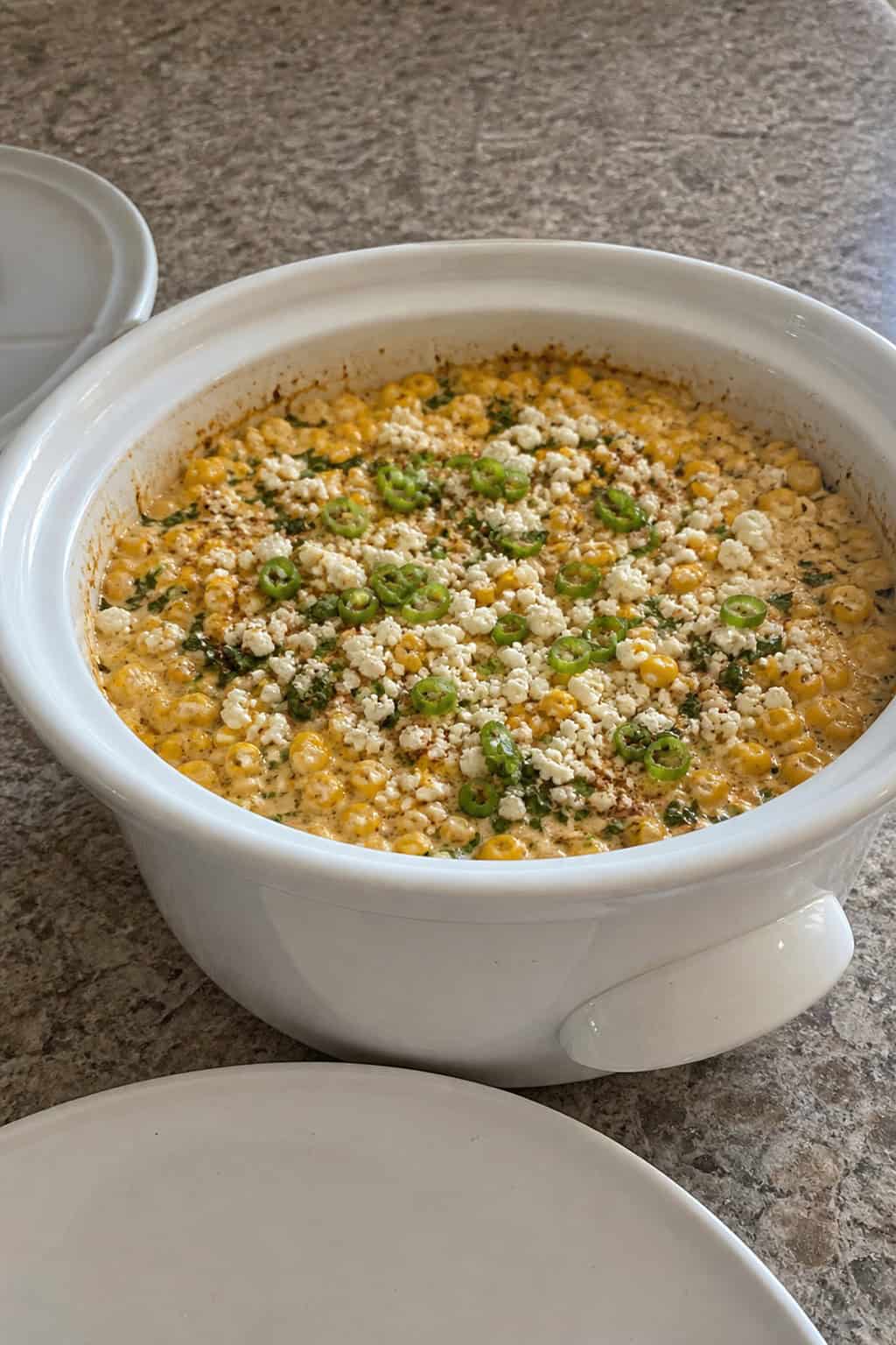 A crockpot filled with creamy Mexican street corn casserole on a wooden table surrounded by fresh corn, lime wedges, and cheese.