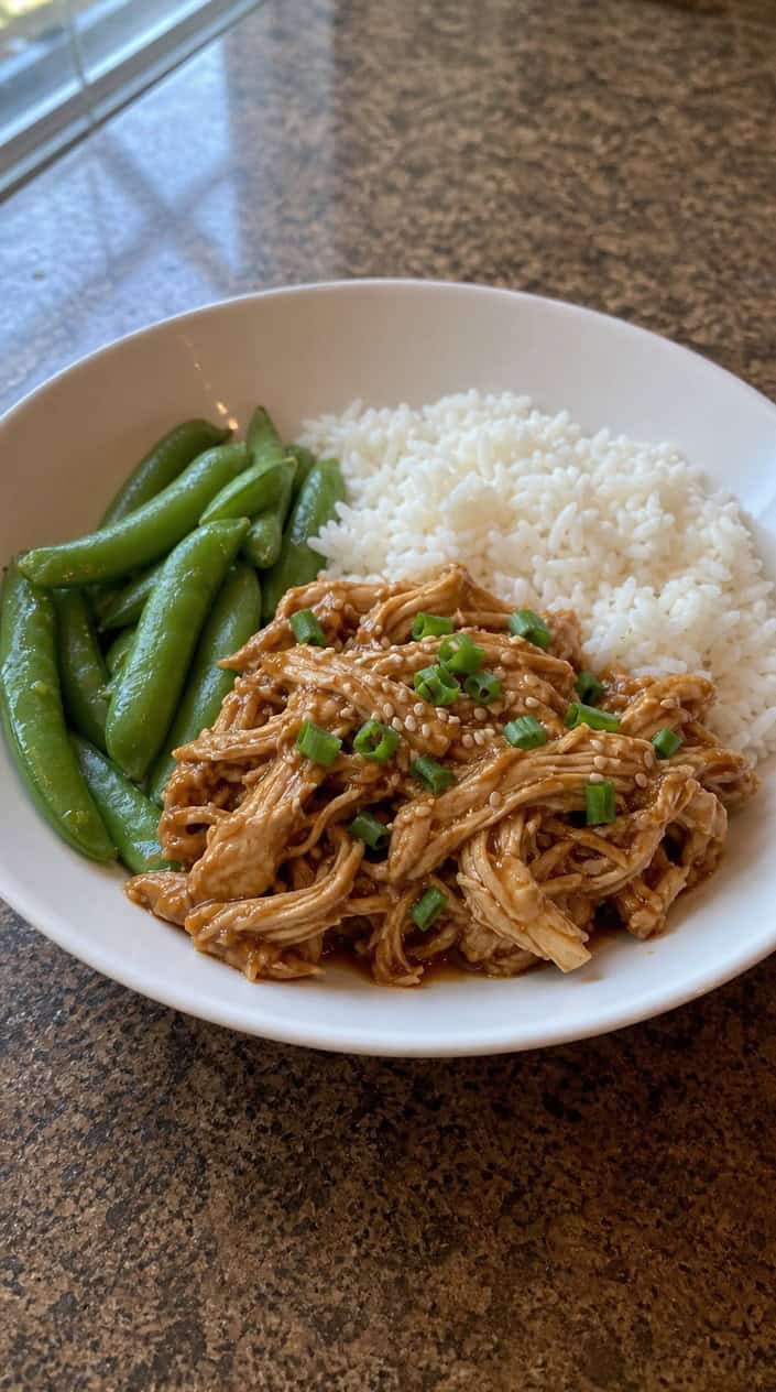 A bowl of glossy orange-ginger shredded chicken over steamed white rice with bright green snap peas, topped with sesame seeds and sliced green onion on a wooden table with fresh ingredients around it.