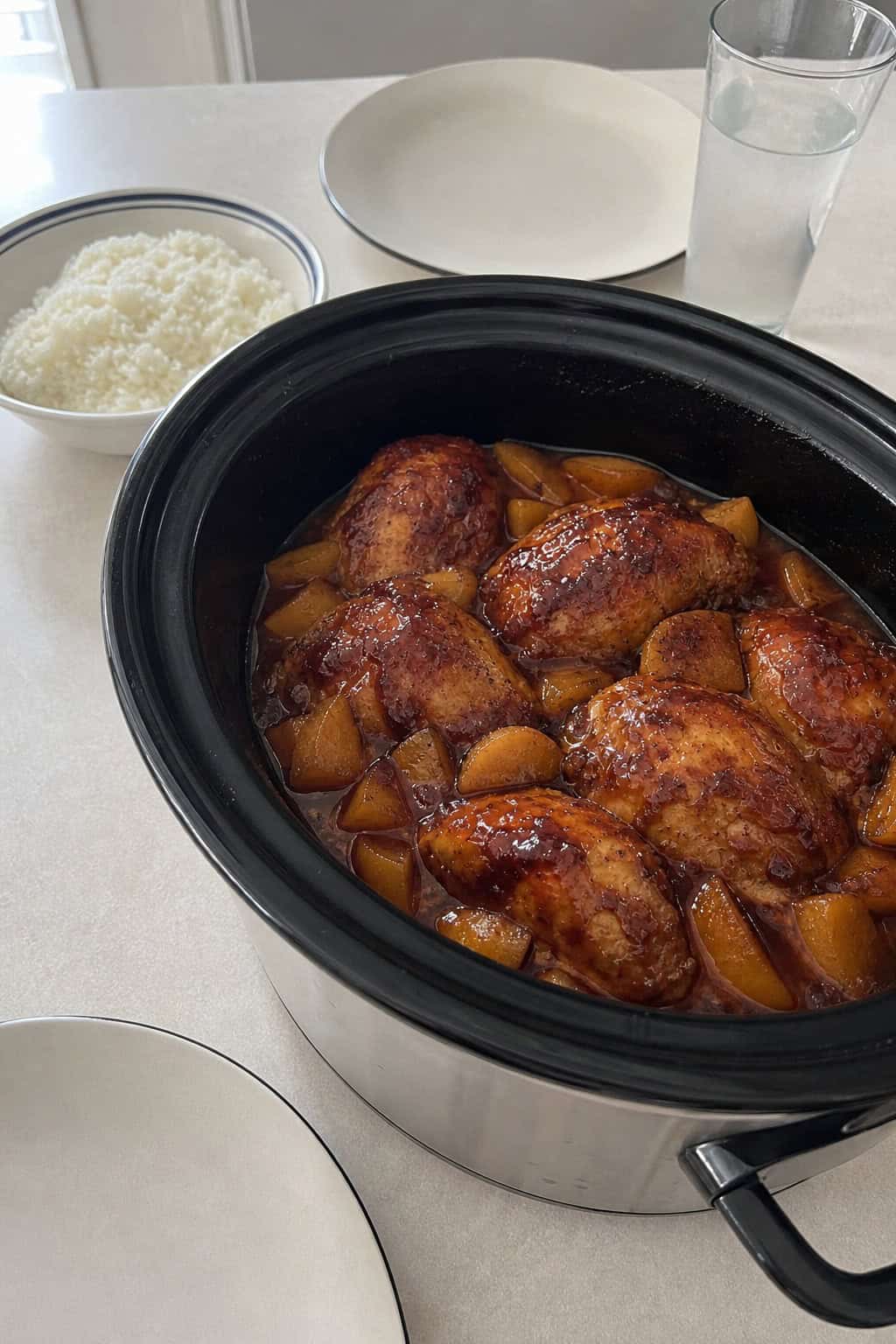 A crockpot filled with peach BBQ chicken surrounded by fresh peaches and ingredients on a kitchen counter.