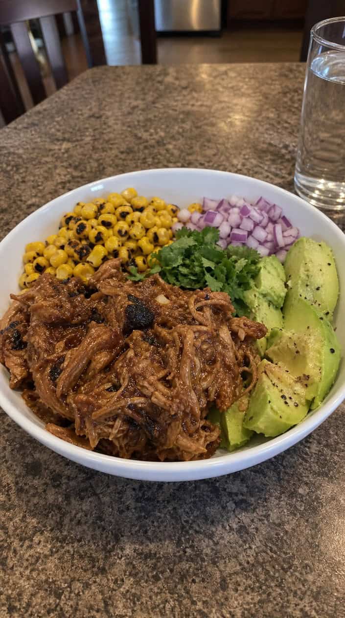 A rustic bowl of pull-apart pollo asado chicken with blistered yellow corn, fanned avocado slices, and a scatter of fresh cilantro on a worn wooden table.