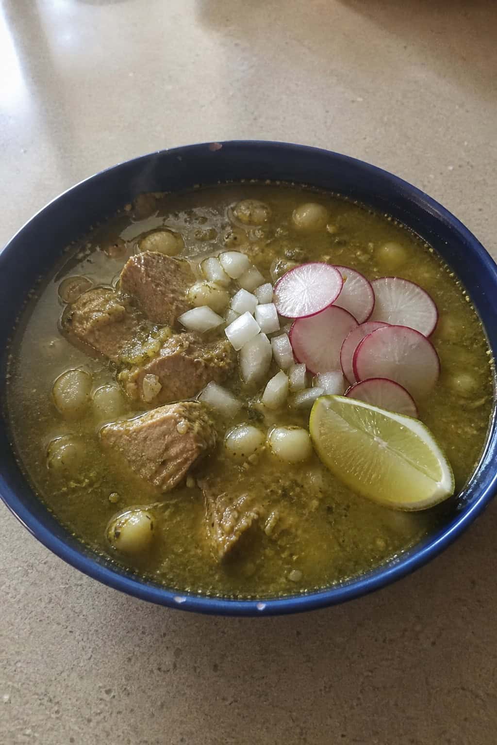 A bowl of green pozole soup with pork, hominy, and fresh garnishes on a wooden table.