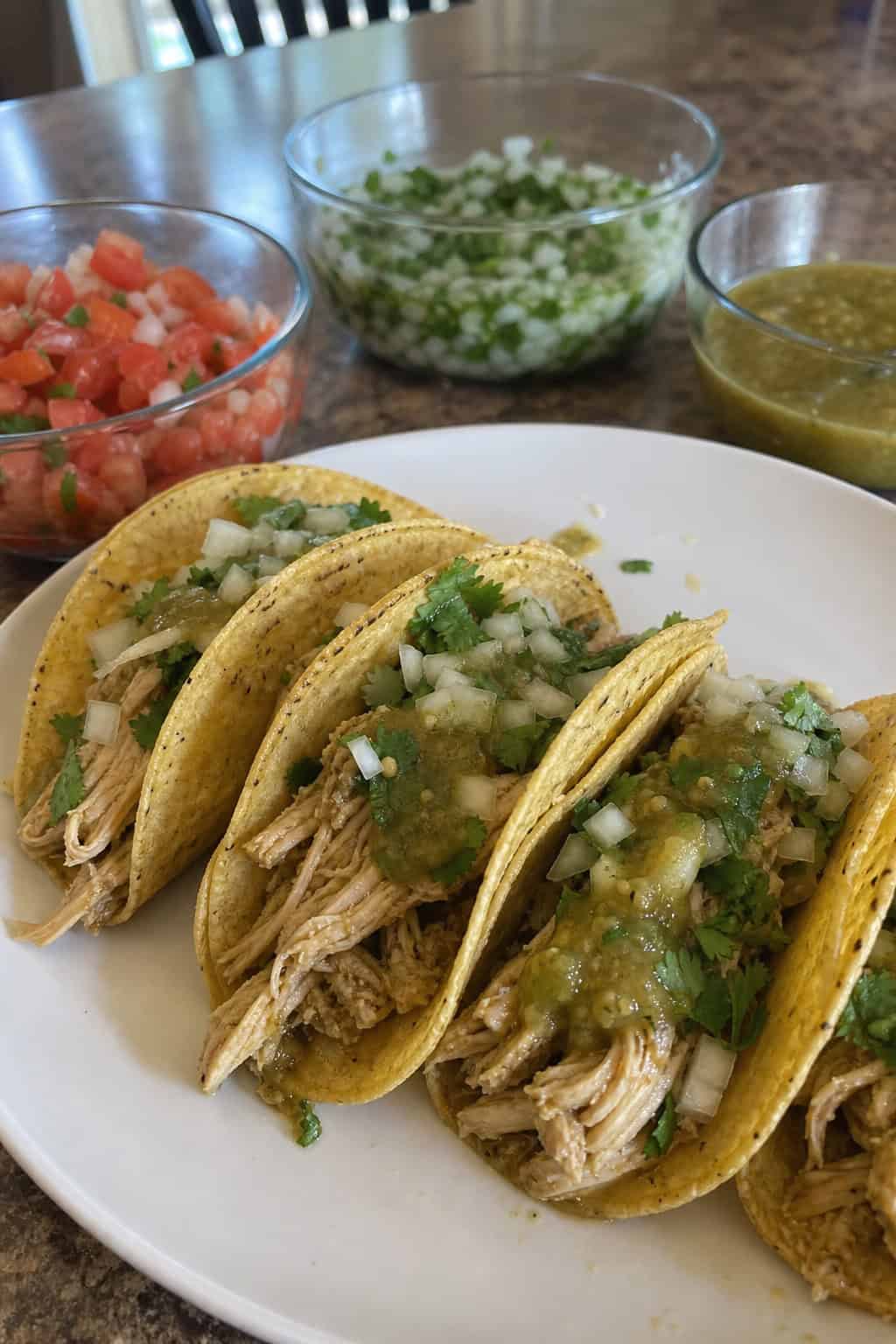 A plate of crockpot salsa verde chicken tacos with fresh toppings on a wooden table surrounded by summer ingredients.