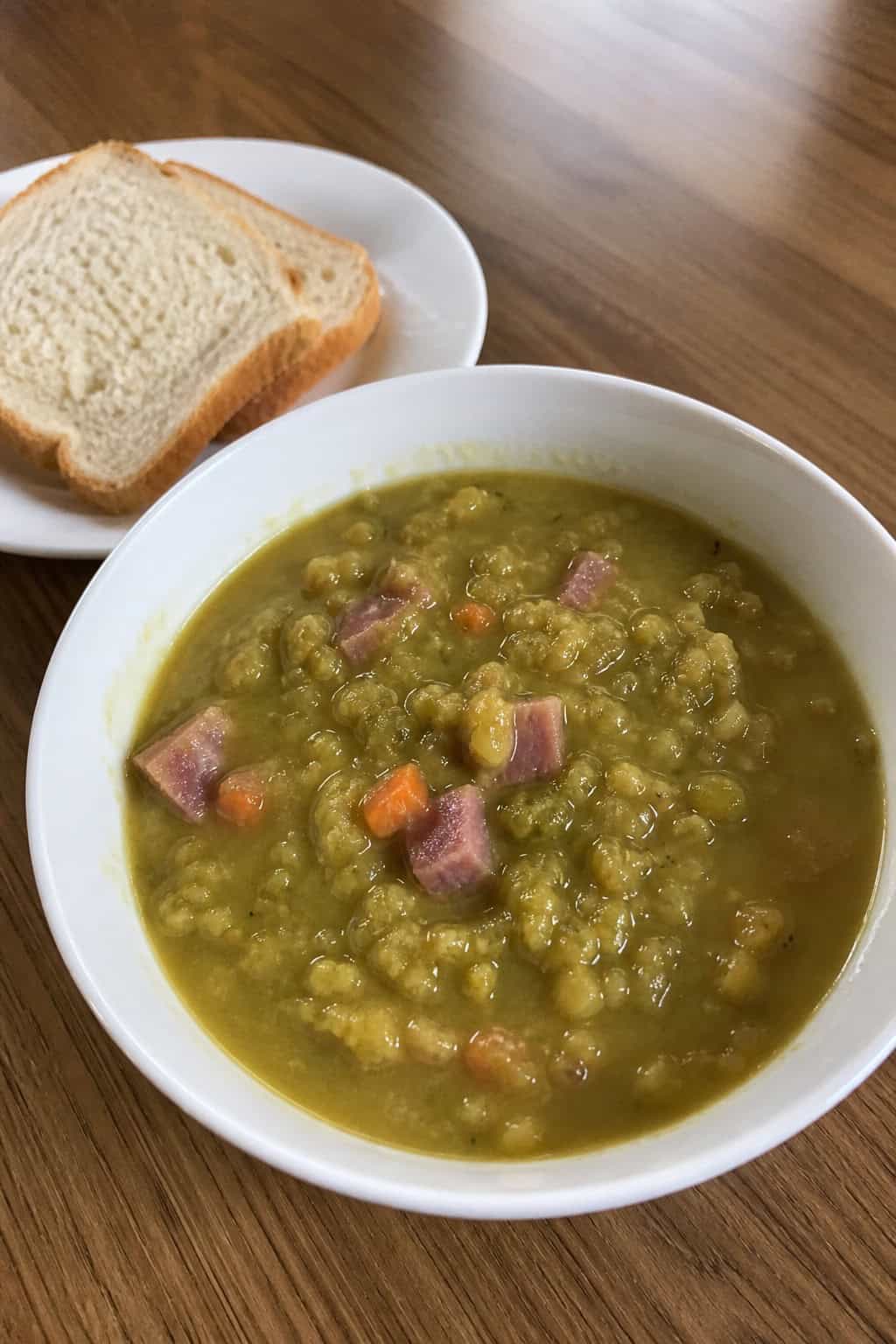 A bowl of split pea and ham soup on a wooden table with fresh ingredients and bread nearby.