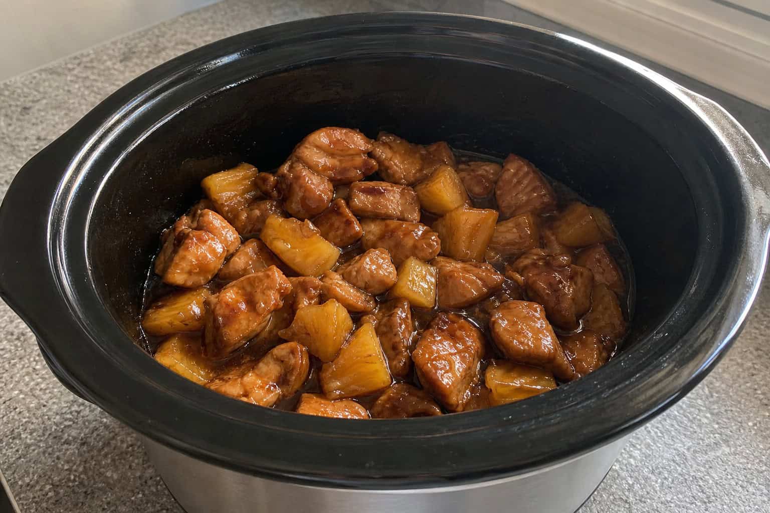 A crockpot filled with teriyaki chicken and pineapple chunks, garnished with green onions on a kitchen countertop with fresh ingredients around.