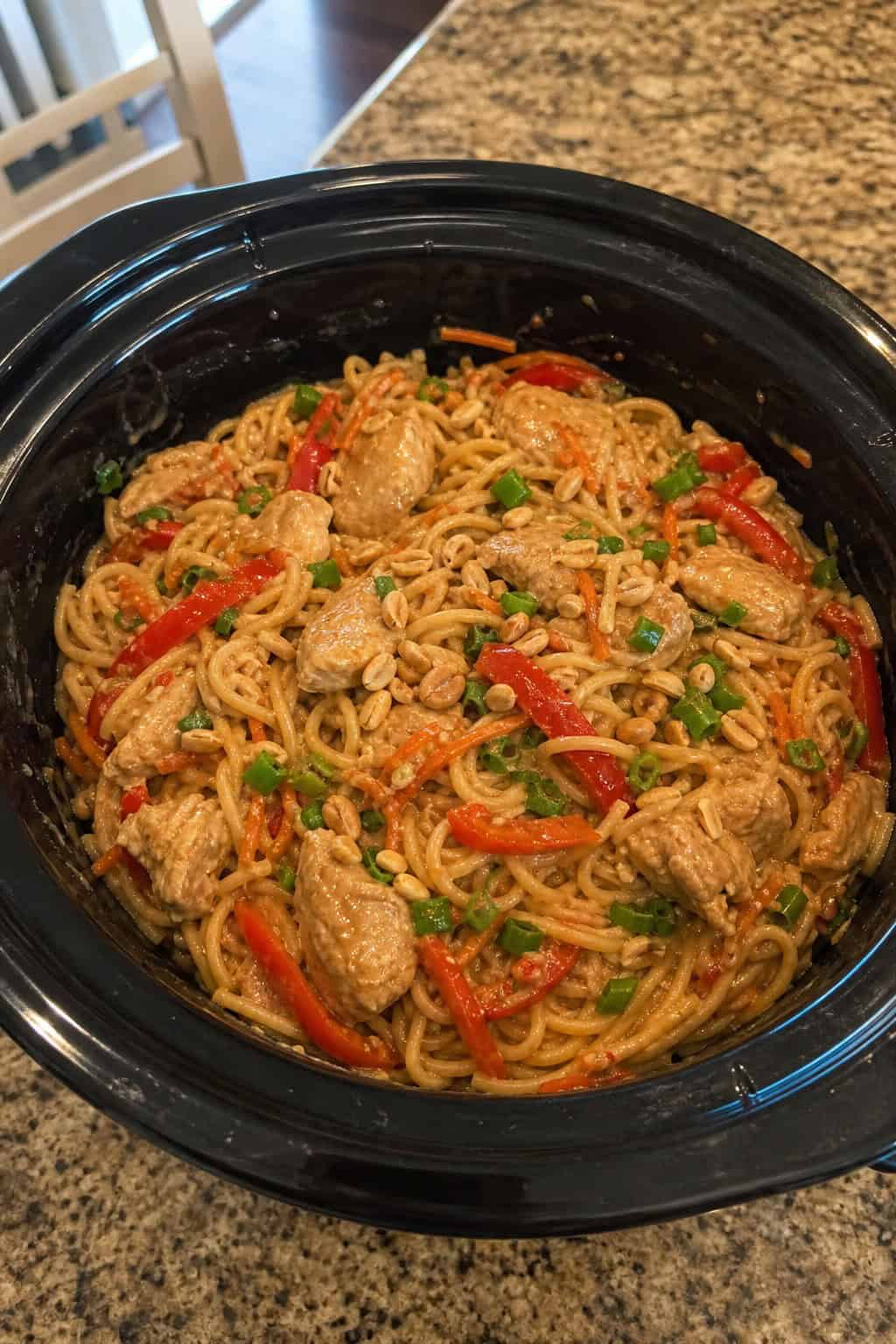 A crockpot filled with Thai peanut chicken noodles, surrounded by fresh ingredients on a kitchen countertop.