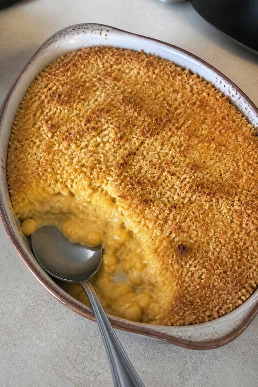 A corn casserole with a deeply golden, rough-textured panko breadcrumb topping in a white ceramic dish on a wooden table, with a large serving spoon resting on the side.