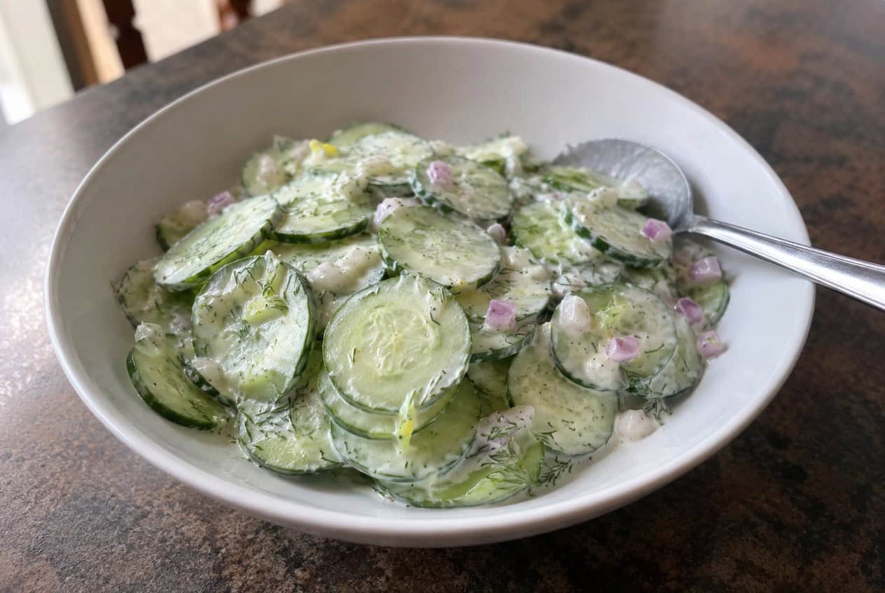 A white ceramic bowl filled with thin cucumber rounds coated in a creamy white yogurt dressing flecked with green dill, with lemon halves and fresh dill sprigs arranged around the bowl on a wooden surface.