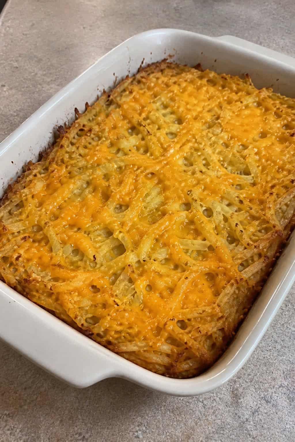 A baked hashbrown casserole with a lightly browned top and visible herb flecks, made with vegan cheese, in a rectangular baking dish on a kitchen table with a bowl of fruit and a glass of juice nearby.