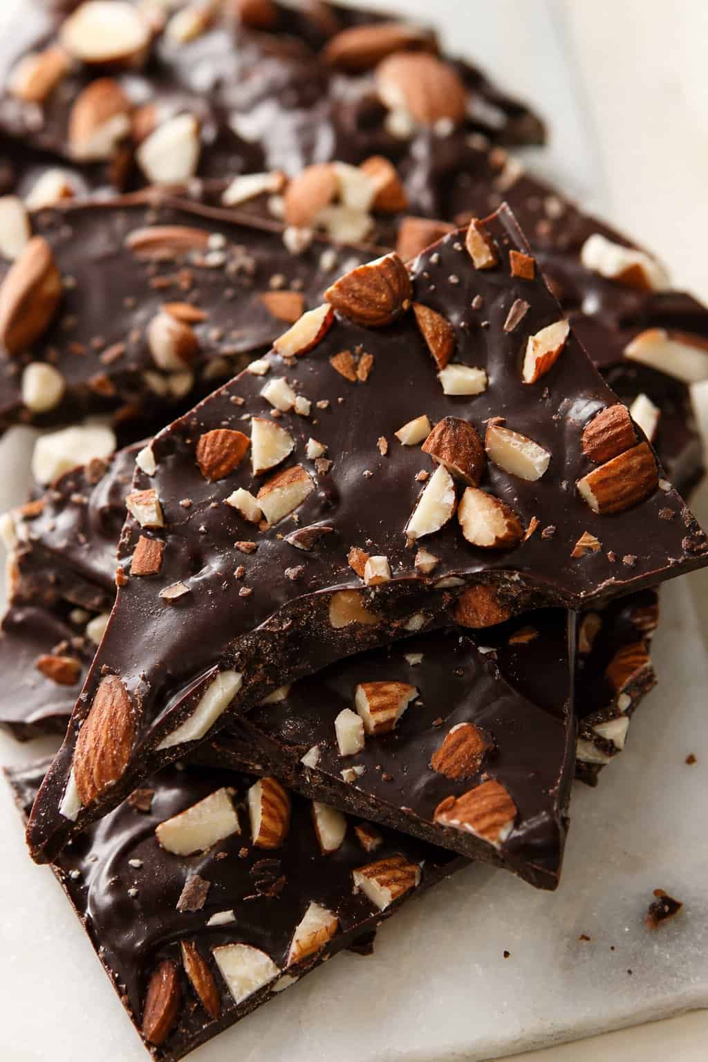 Jagged shards of dark chocolate almond bark on a sheet of white parchment paper, with coarsely chopped toasted almonds pressed into the surface and a few flakes of sea salt visible on top.