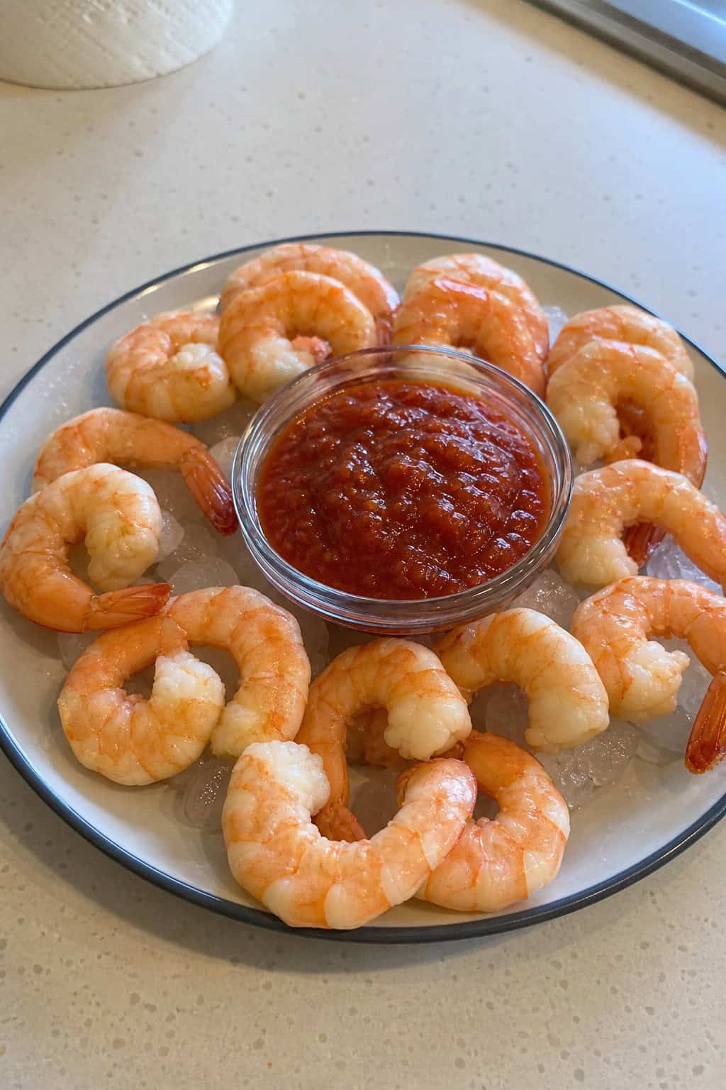 A platter of large tails-on shrimp arranged around a bowl of red cocktail sauce with lemon wedges and parsley on a white surface.