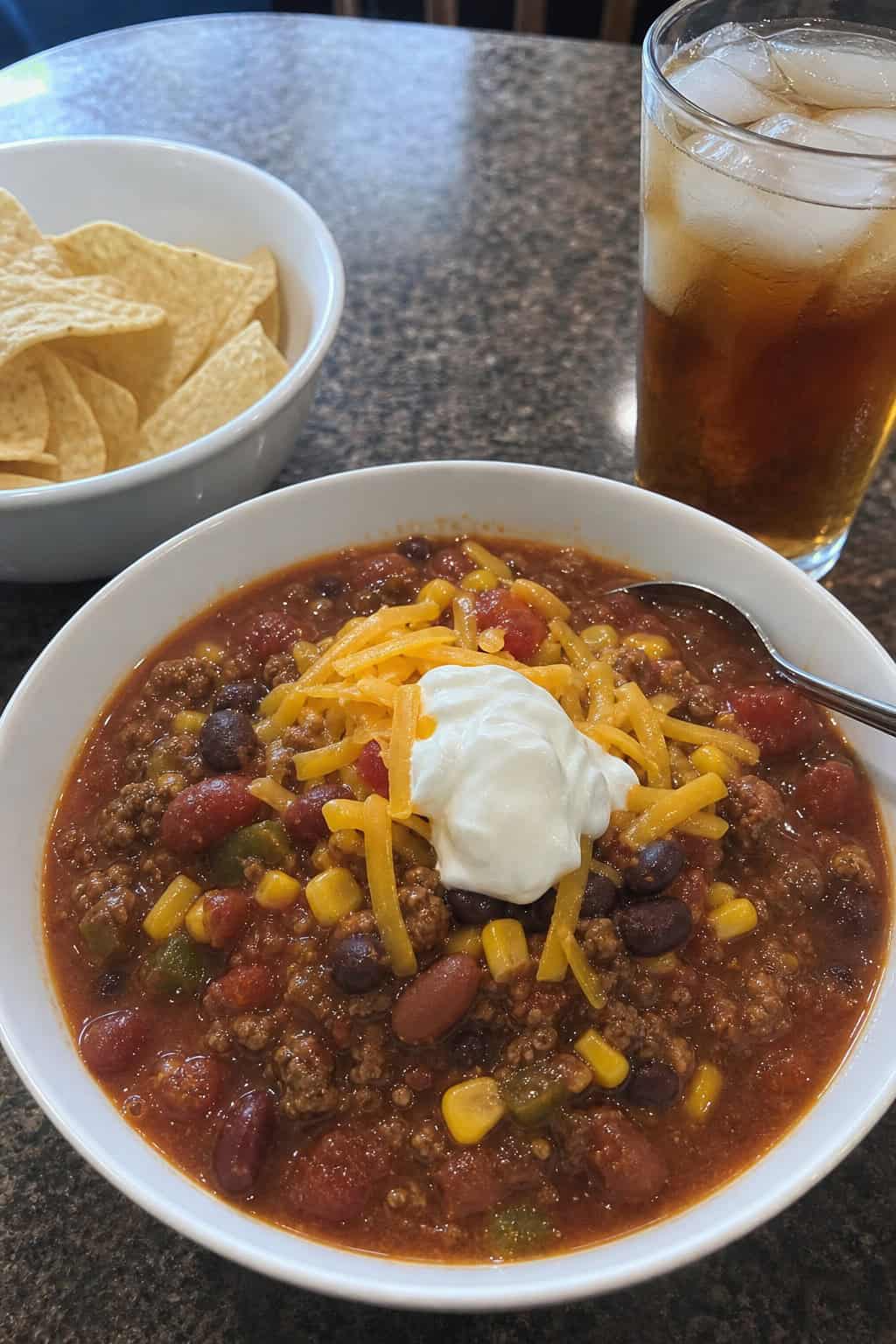 A bowl of taco chili with beans, corn, and cheese on a wooden table, surrounded by tortilla chips and a glass of iced tea.