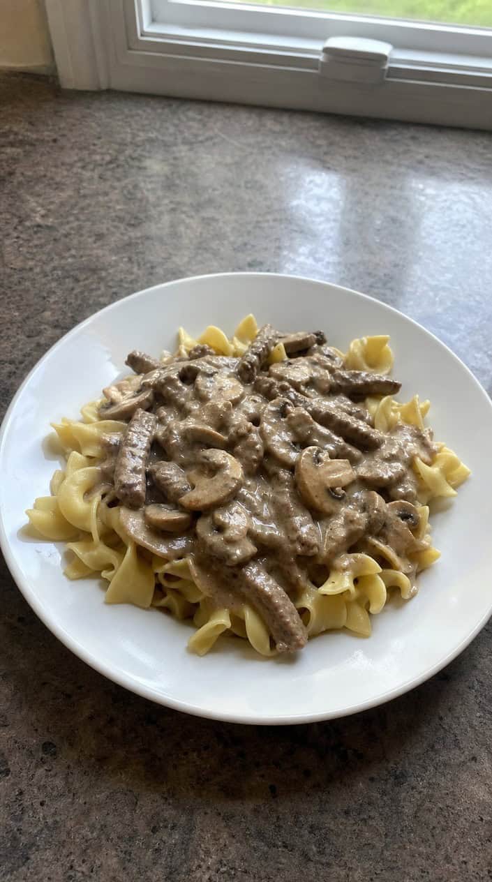 A bowl of creamy beef stroganoff with mushrooms and parsley served over egg noodles on a wooden table.