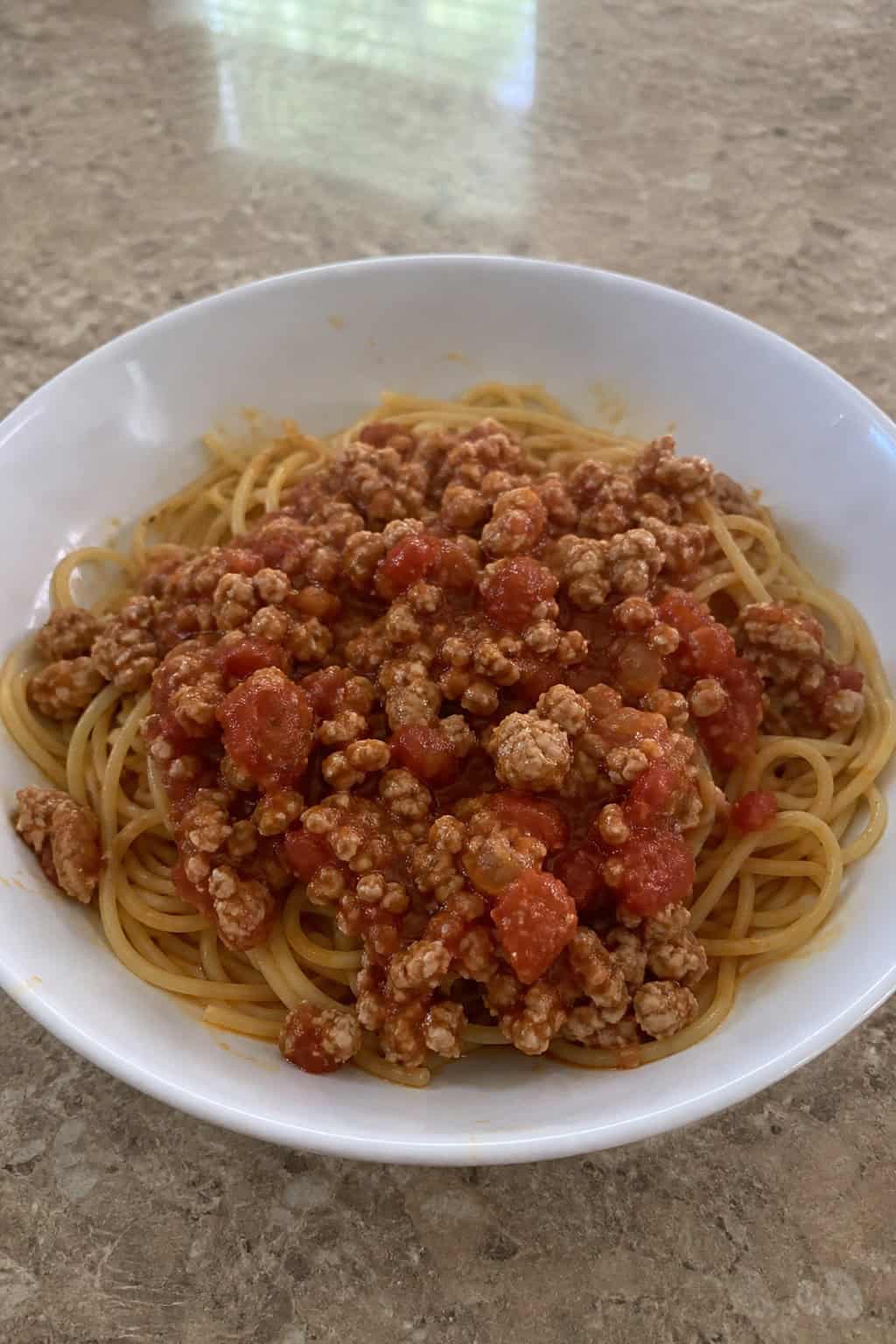 A bowl of minced chicken bolognese served over pasta on a wooden table with fresh ingredients in the background.