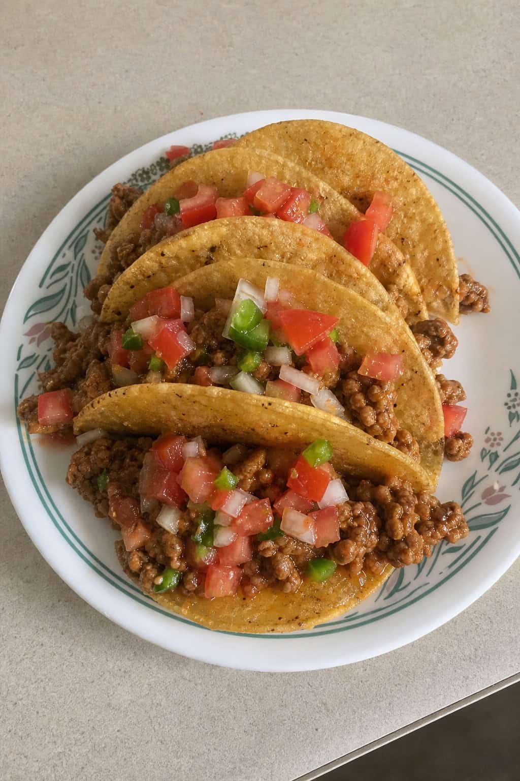 A plate of minced chicken tacos topped with fresh pico de gallo on a wooden table with a bowl of salsa and lime wedges nearby.