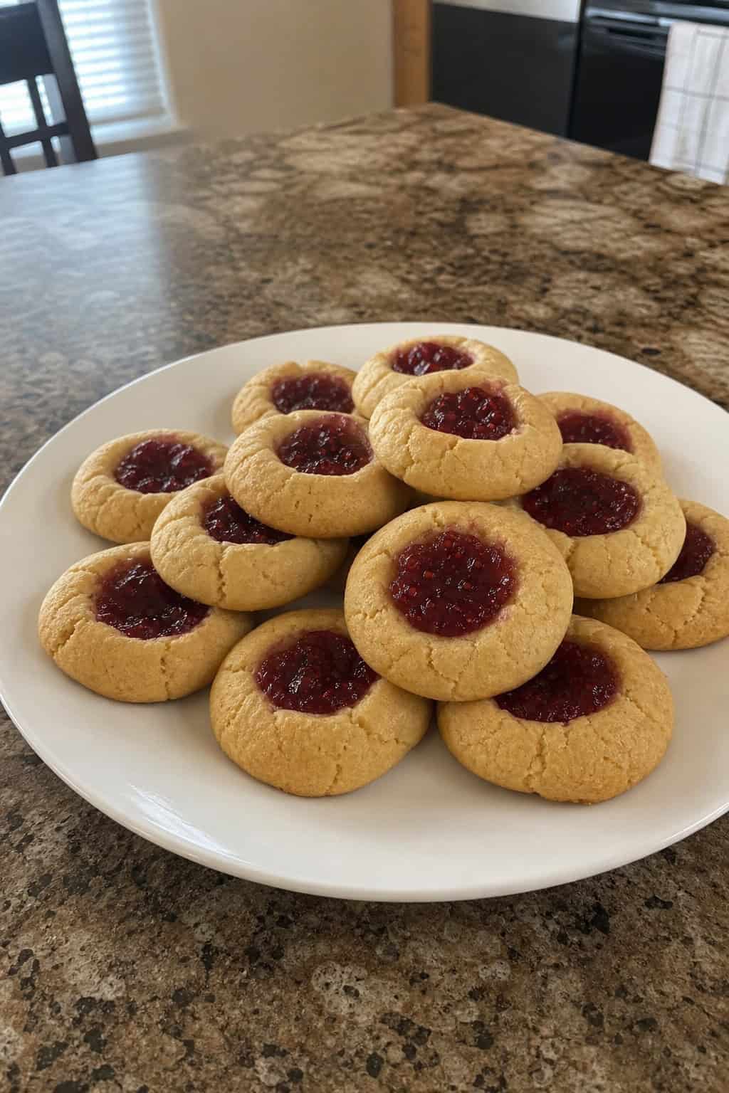 A plate of golden thumbprint jam cookies on a wooden table with jam and baking ingredients nearby.
