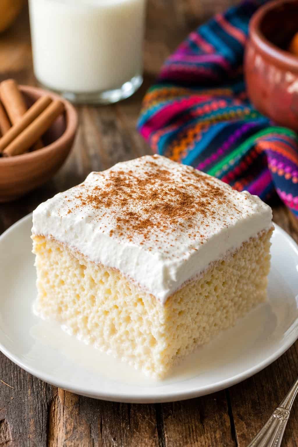 A slice of moist tres leches cake topped with whipped cream and cinnamon on a white plate on a wooden table.