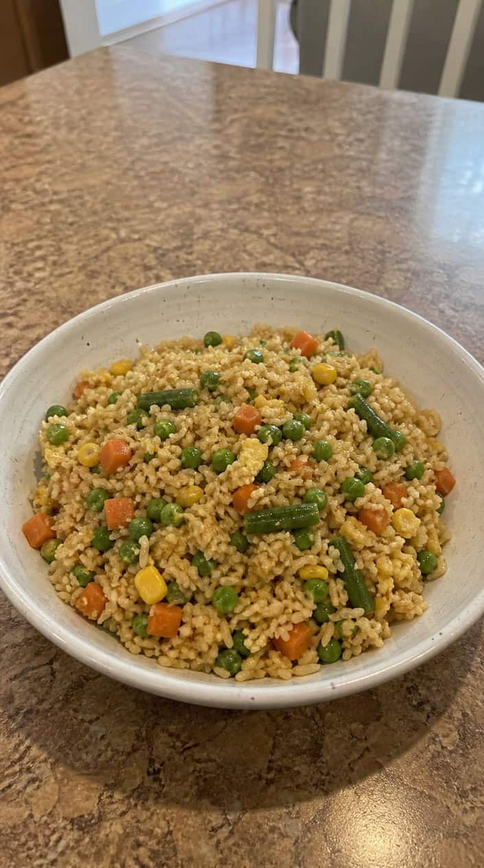 A ceramic bowl of golden egg fried rice with visible flecks of scrambled egg, diced green vegetable, and shredded carrot, garnished with sliced green onion, photographed on a dark wood table.