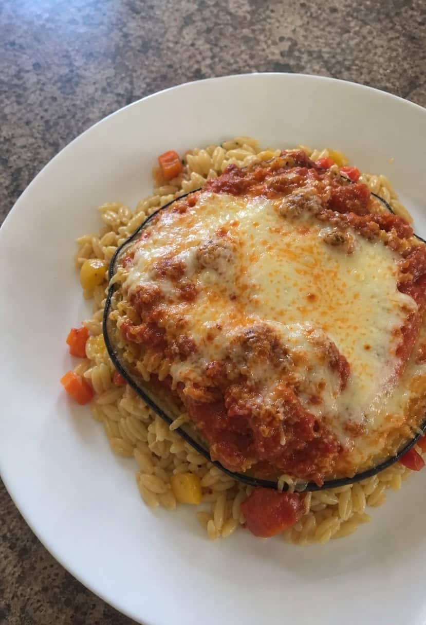 A steaming hot plate of eggplant parmesan surrounded by colorful ingredients and herbs on a rustic wooden table