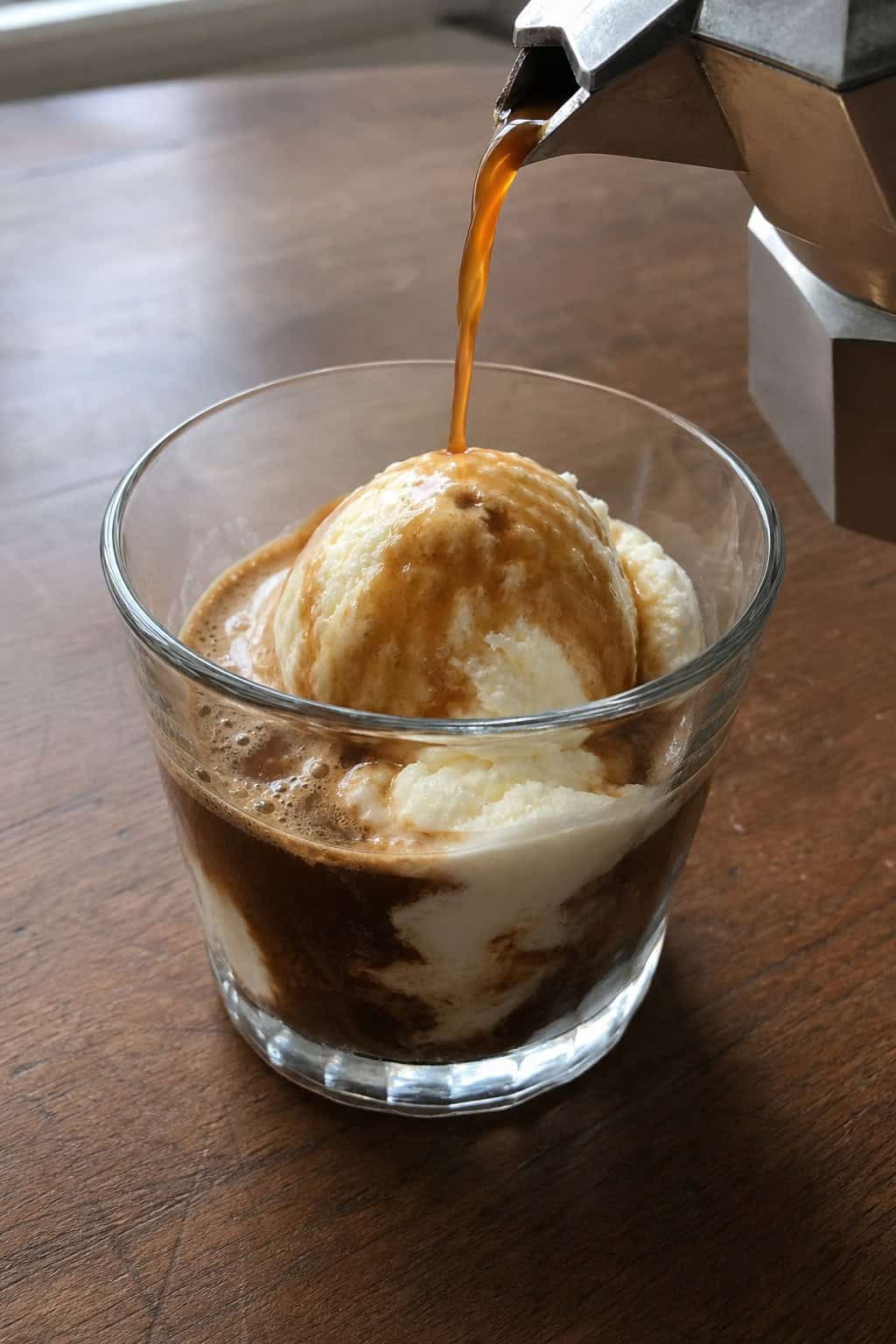 A glass cup of espresso being poured over a scoop of vanilla ice cream on a wooden table.