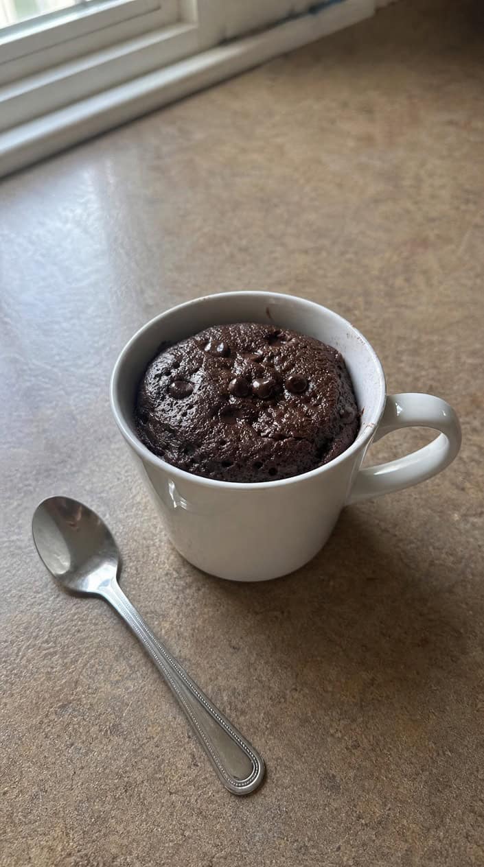 A white ceramic mug containing a dark chocolate mug cake with visible mini chocolate chips at the surface, set on a light wood countertop with whole coffee beans scattered beside it and a silver spoon resting against the mug.