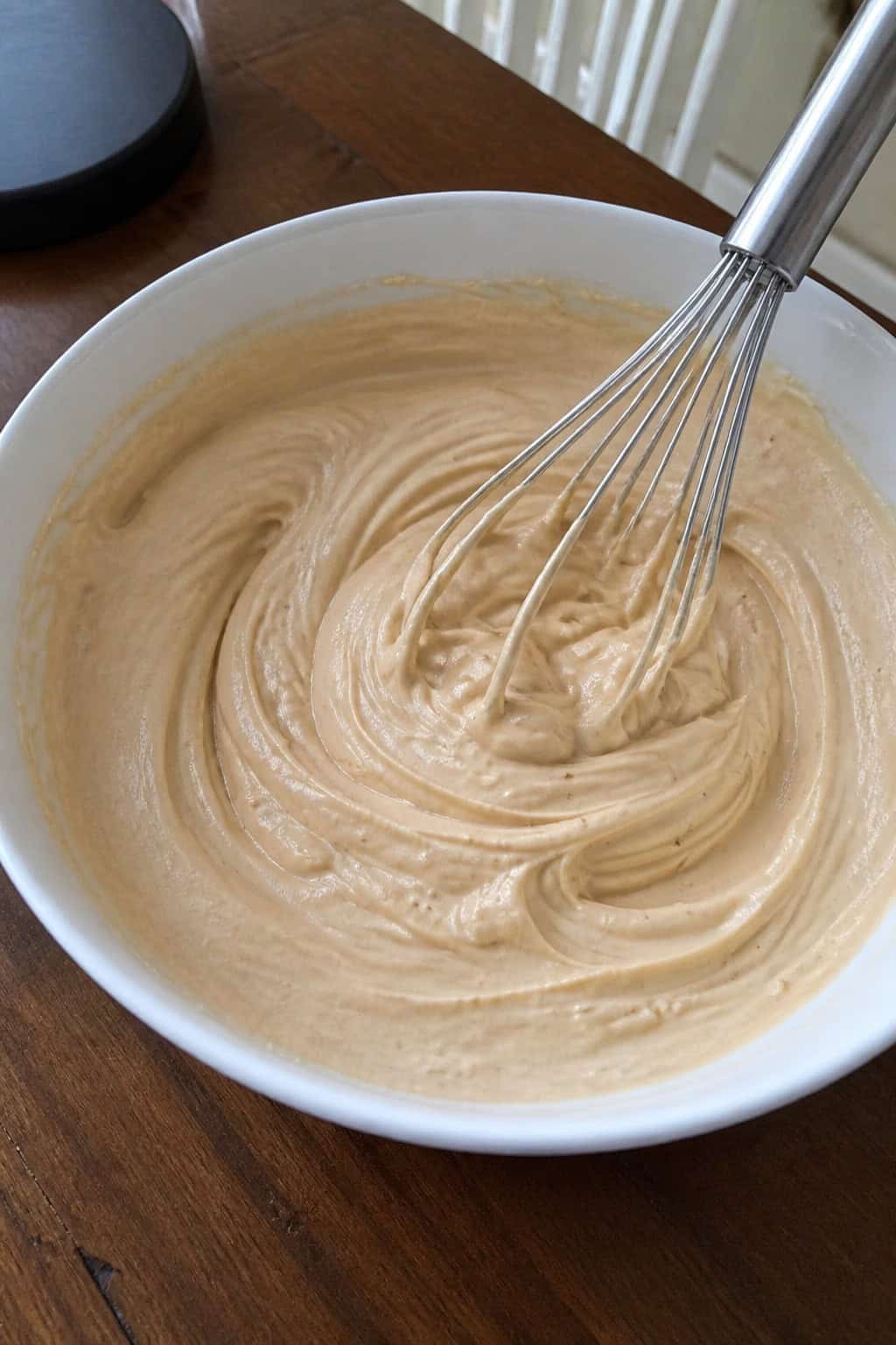 Close-up of creamy espresso-infused cheesecake filling in a glass bowl with a whisk, coffee beans, and an espresso cup on a wooden surface.