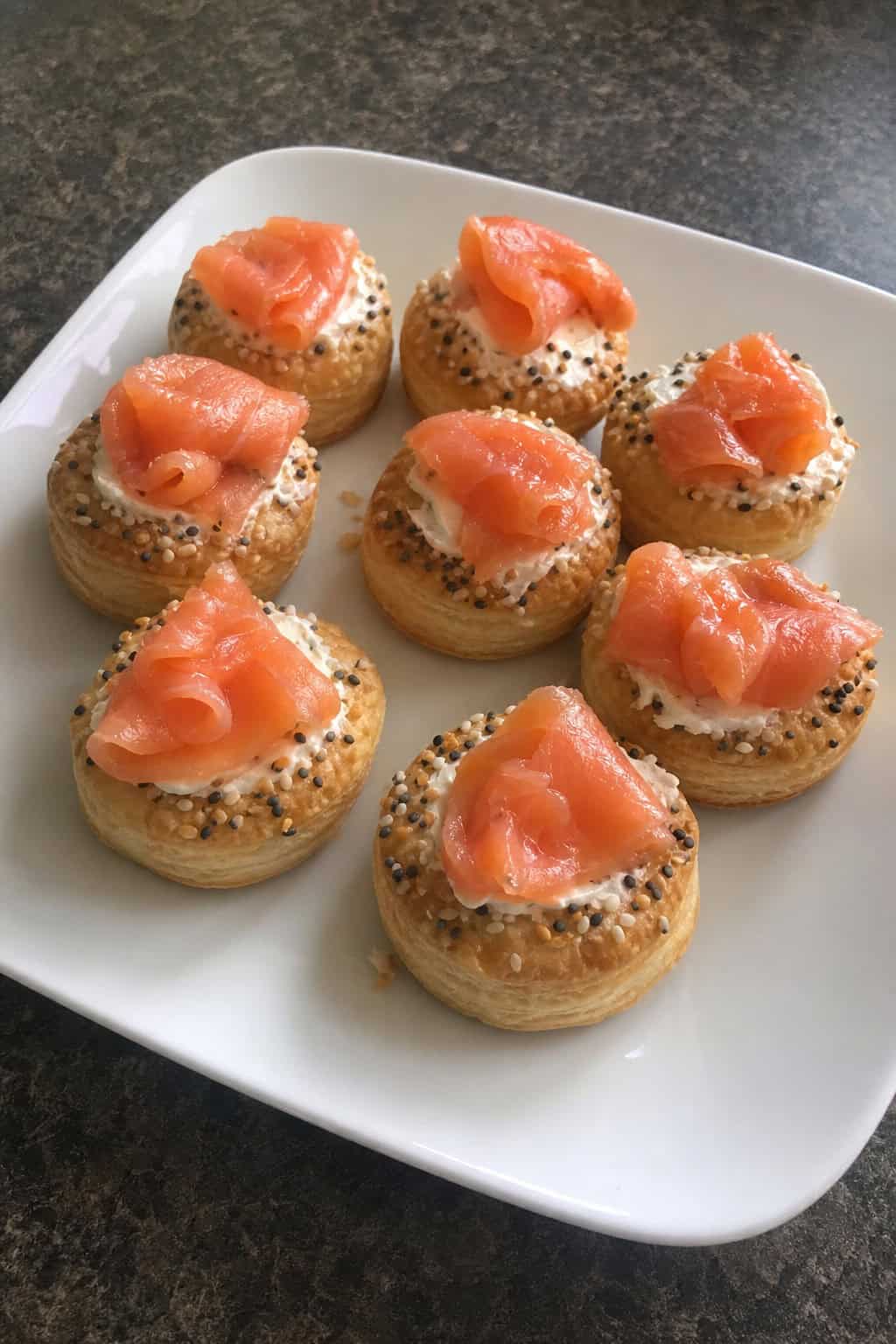 Close-up of bite-sized puff pastry appetizers topped with smoked lox, cream cheese, and fresh herbs arranged on a wooden serving board.
