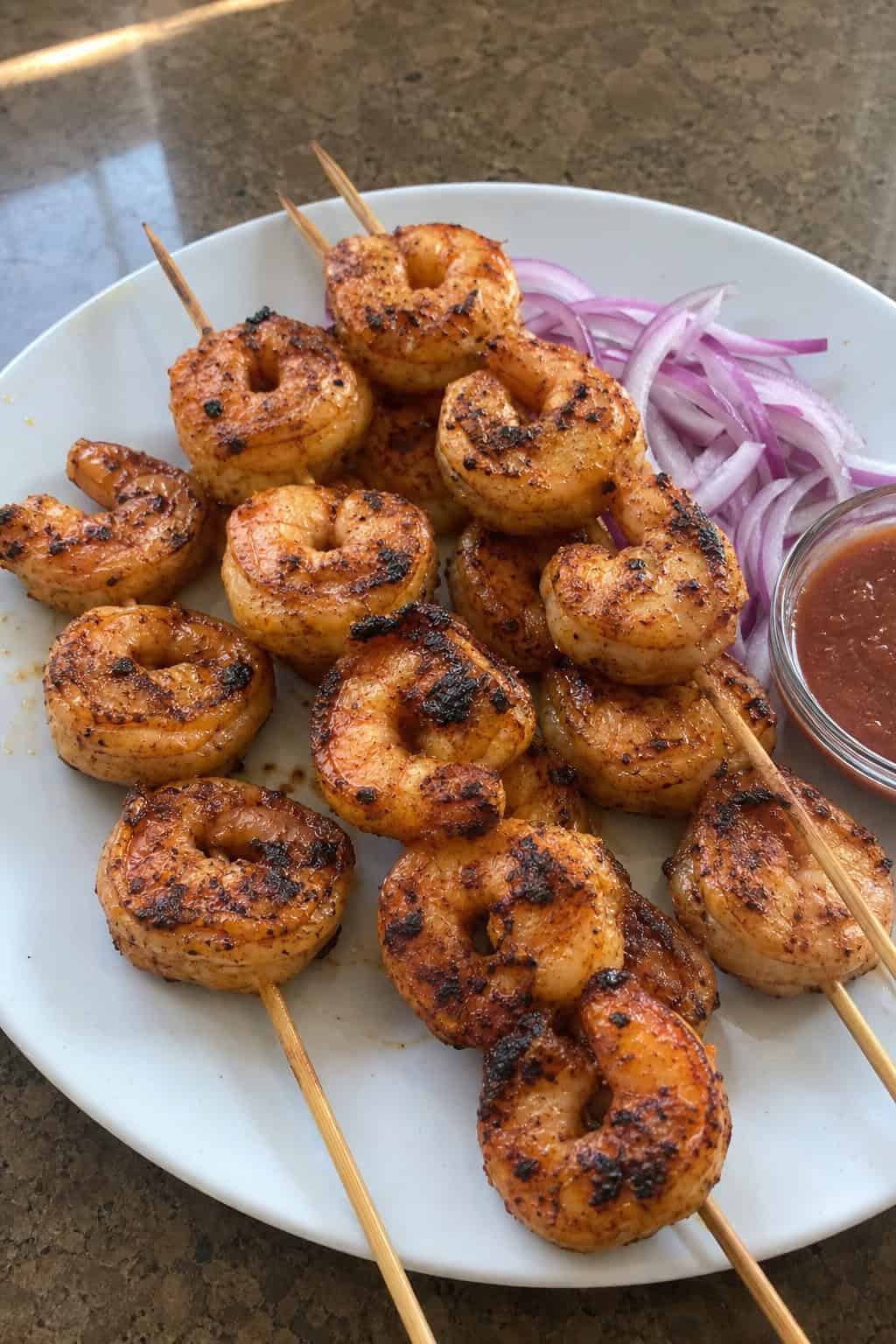 A plate of grilled chipotle-style shrimp skewers garnished with lime wedges, cilantro, and red onions on a wooden table.