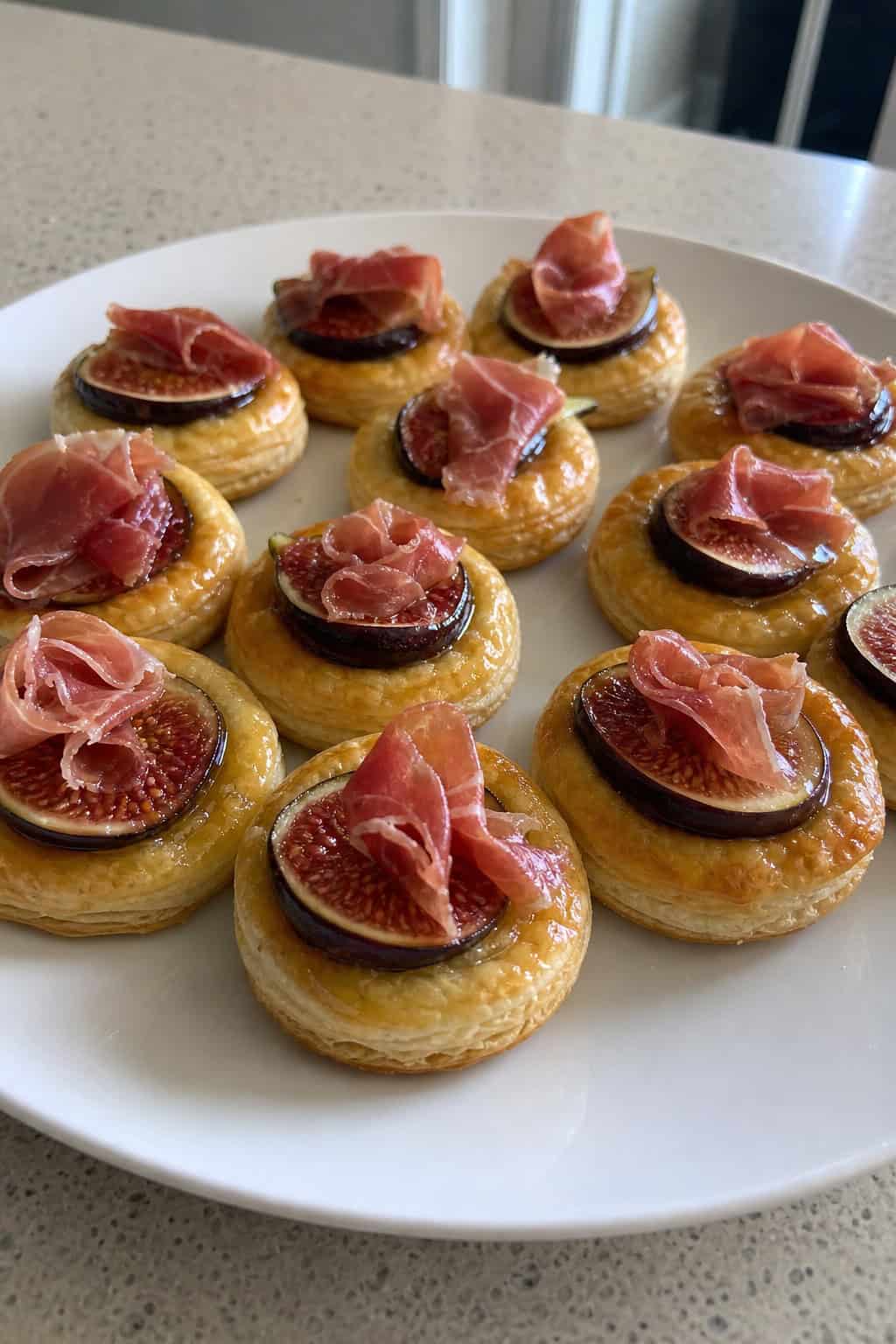 A platter of golden puff pastry rounds topped with slices of fig and prosciutto, garnished with herbs, on a wooden table.