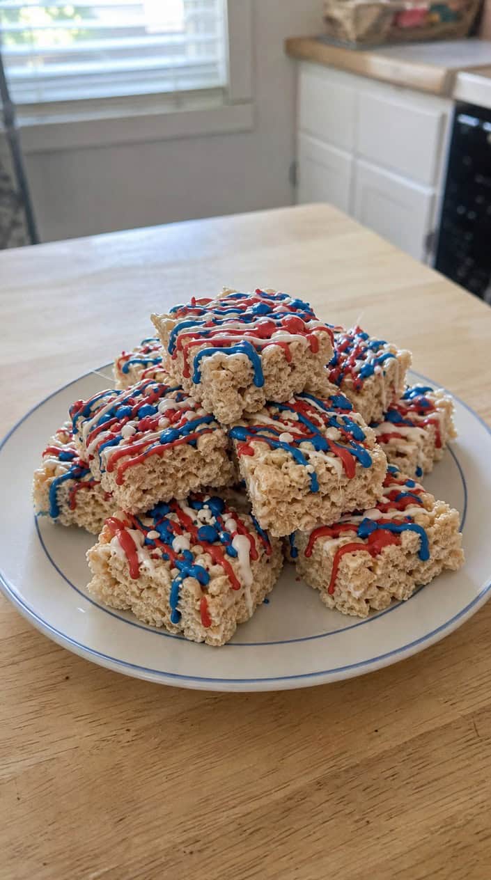 A plate of red, white, and blue drizzled Rice Krispie treats on a wooden table with small American flags and sparklers in the background.