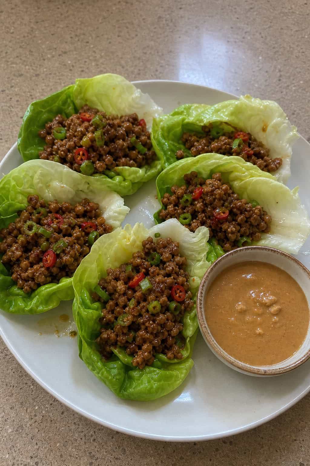 A platter of minced chicken lettuce cups with peanut sauce in a small bowl, surrounded by fresh herbs and ingredients on a wooden surface.