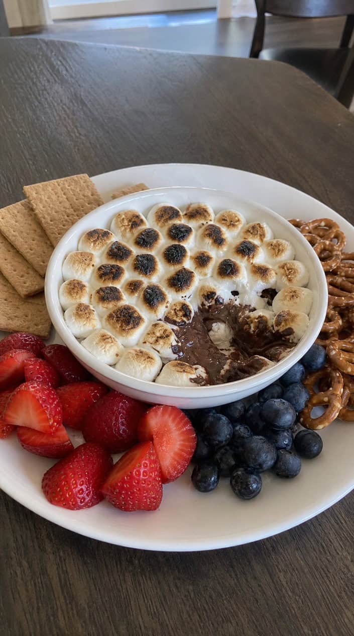 A bowl of s'mores dip topped with toasted marshmallows surrounded by fresh strawberries, blueberries, and white raspberries on a wooden board with small American flags nearby.