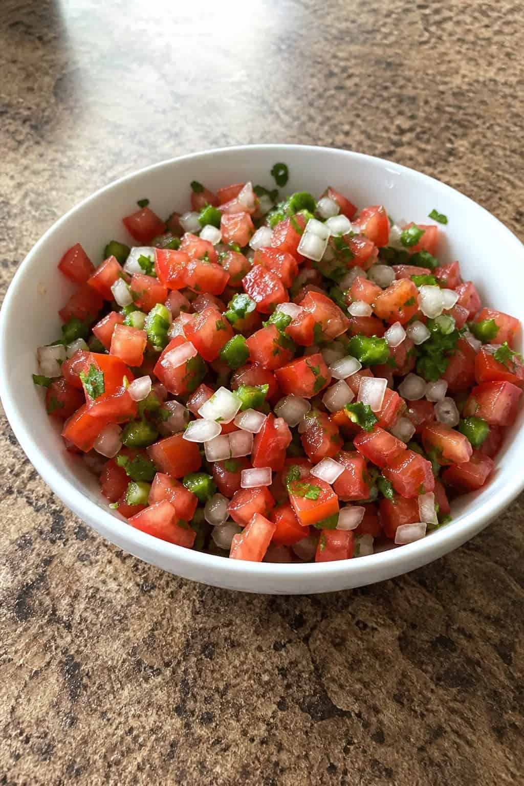 18 Chipotle Copycat Recipes to Make at Home 14 Close-up of a bowl of fresh pico de gallo salsa with tomatoes, onions, cilantro, and jalapeños on a wooden table surrounded by fresh ingredients.