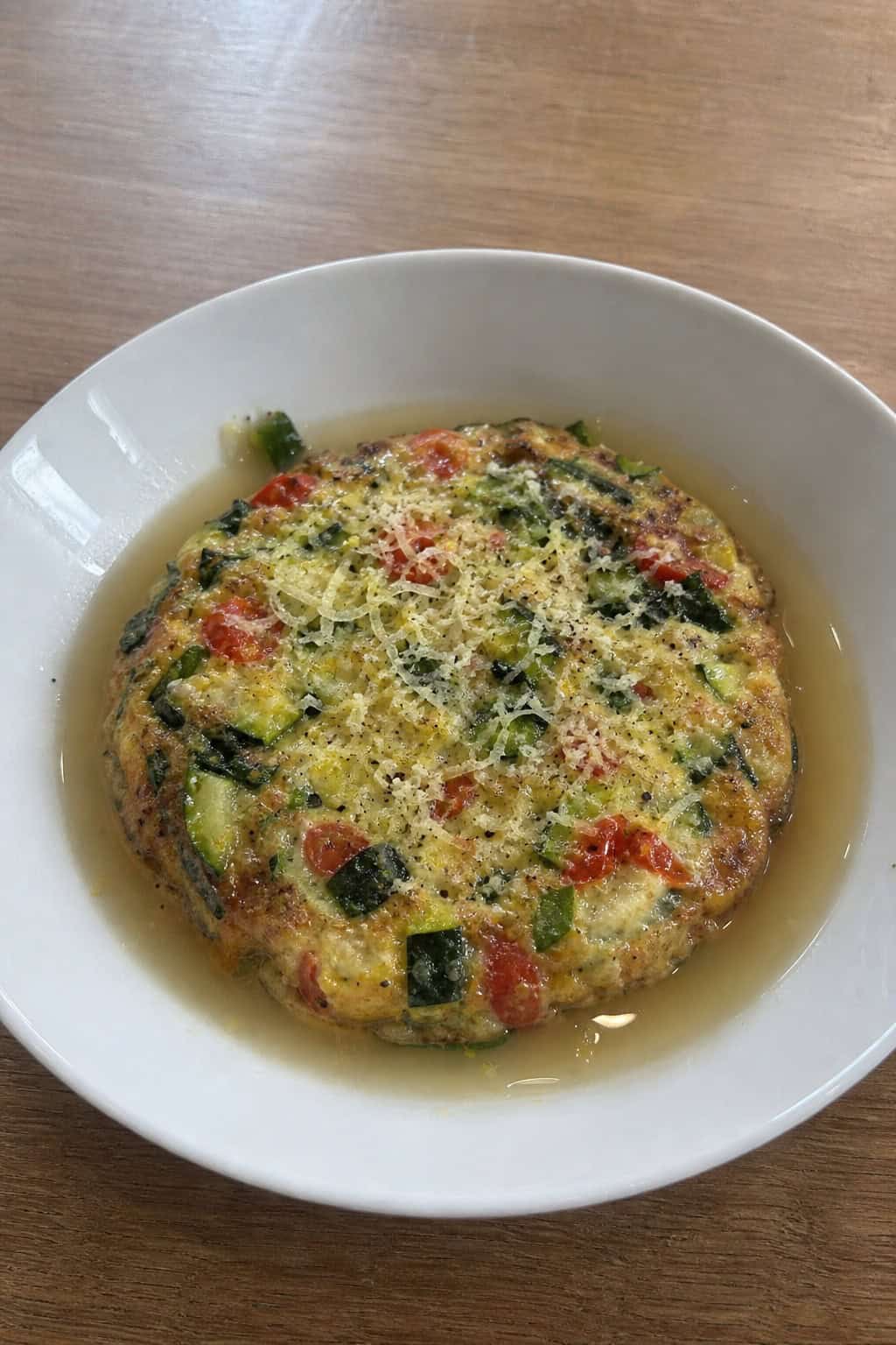 A bowl of vegetable frittata in broth topped with grated rind, placed on a wooden table with simple dining utensils.