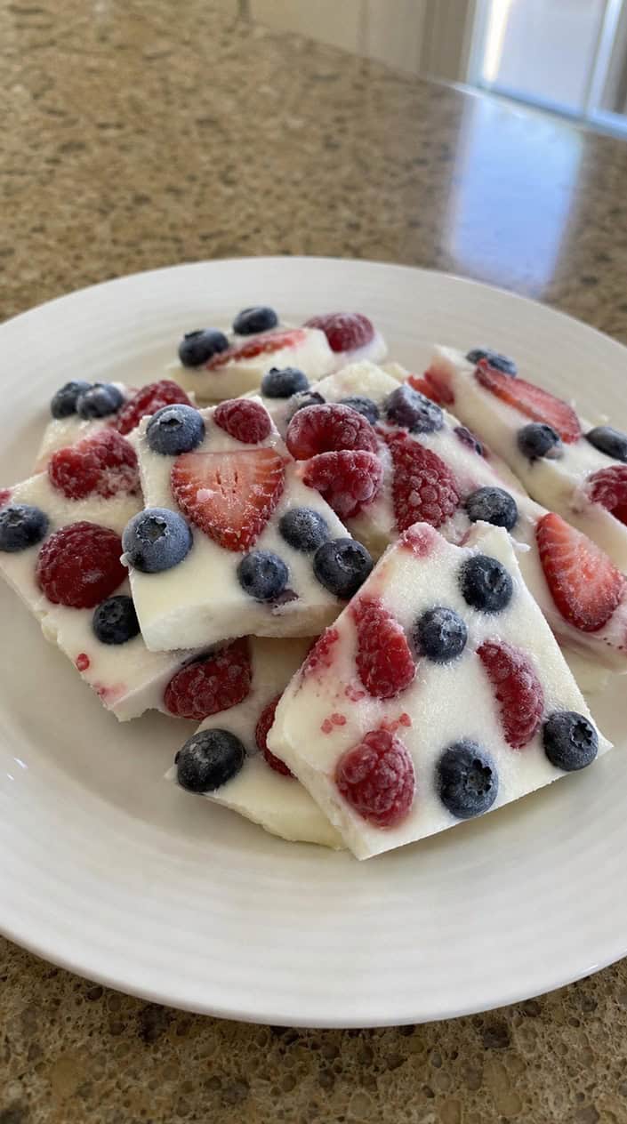 Pieces of frozen Greek yogurt bark topped with fresh berries arranged on a wooden surface with warm sunlight in the background.