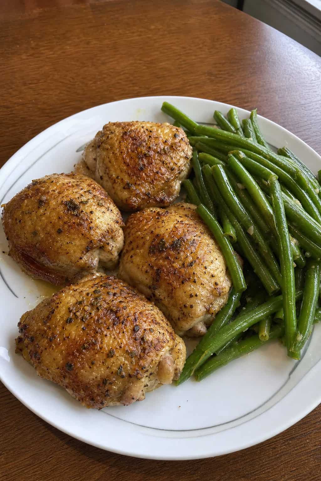 A plate with garlic butter baked chicken thighs and green beans on a wooden table.