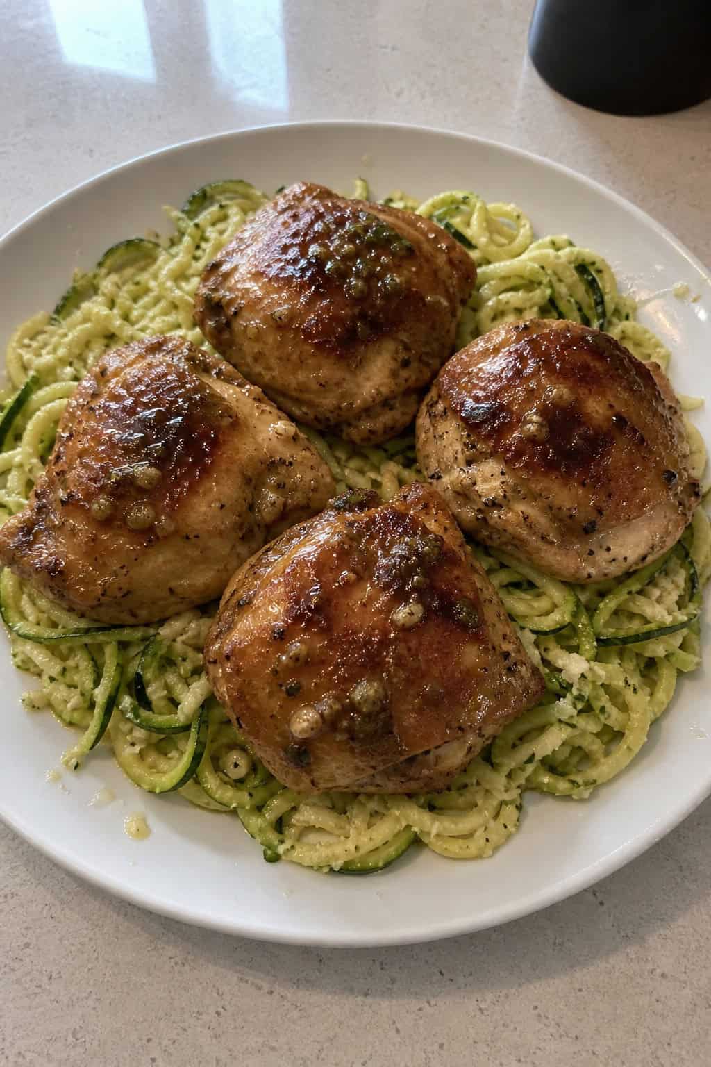 Golden seared chicken thighs plated over a tangle of zucchini noodles glistening with garlic butter, topped with grated Parmesan and a few fresh herb leaves, served on a light-colored plate.