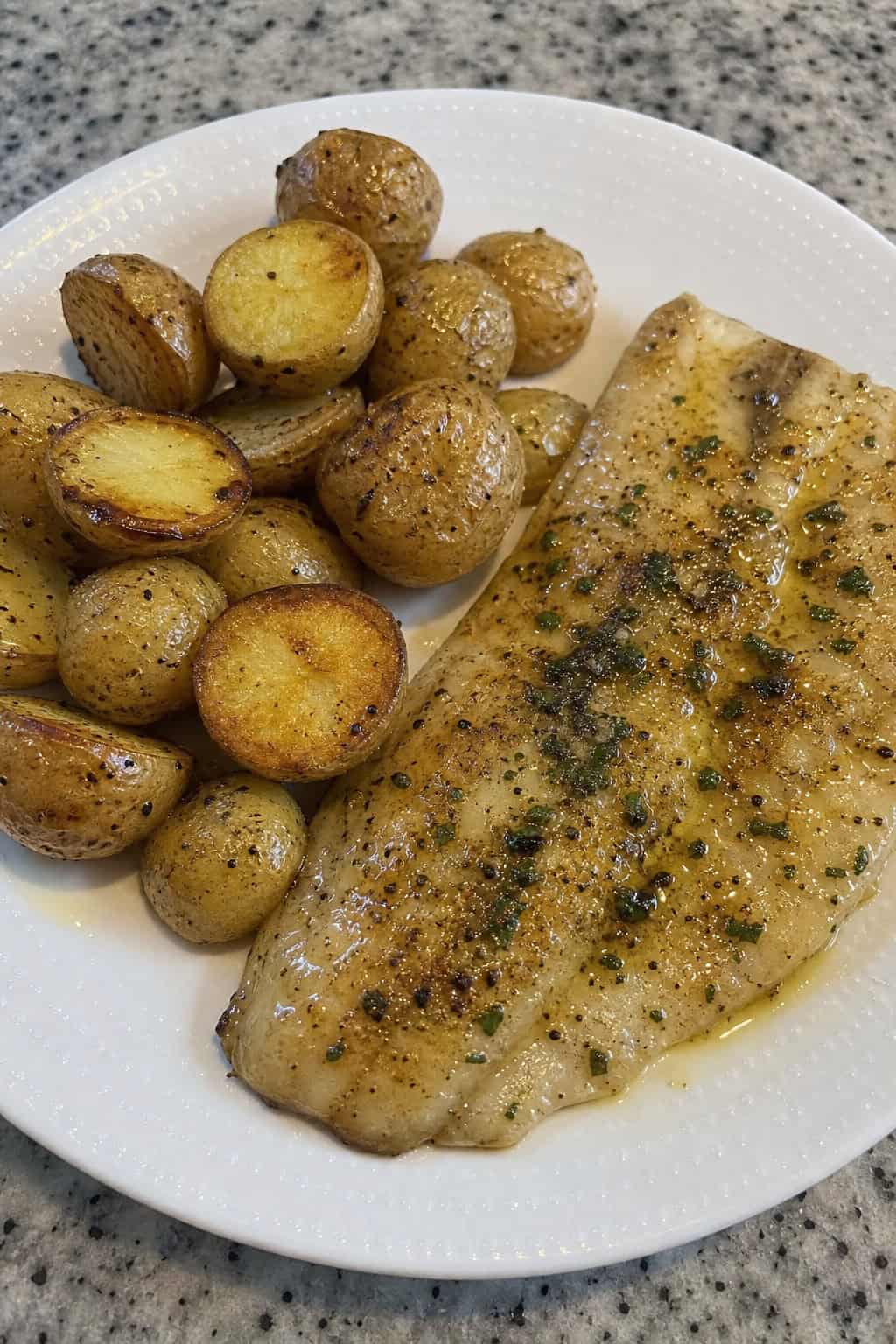 A flaky tilapia fillet with a golden garlic butter crust beside halved roasted baby potatoes garnished with dried parsley on a wooden table.