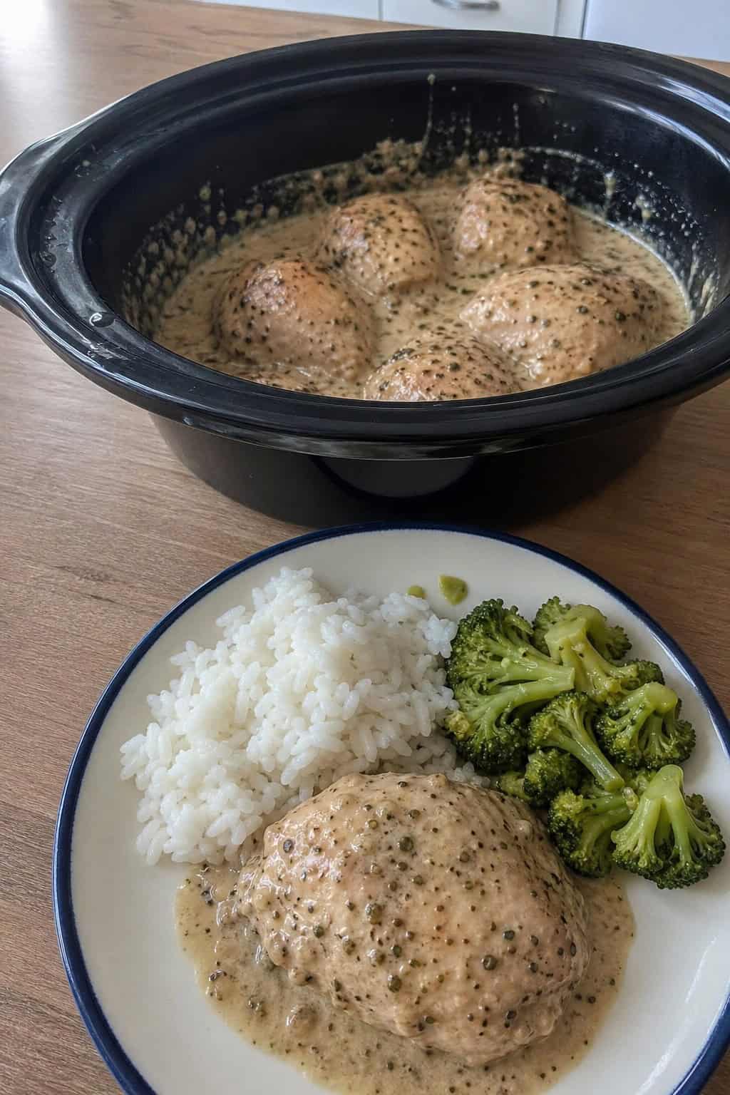 A crockpot filled with garlic parmesan chicken surrounded by fresh garlic, parsley, and parmesan cheese on a kitchen countertop.