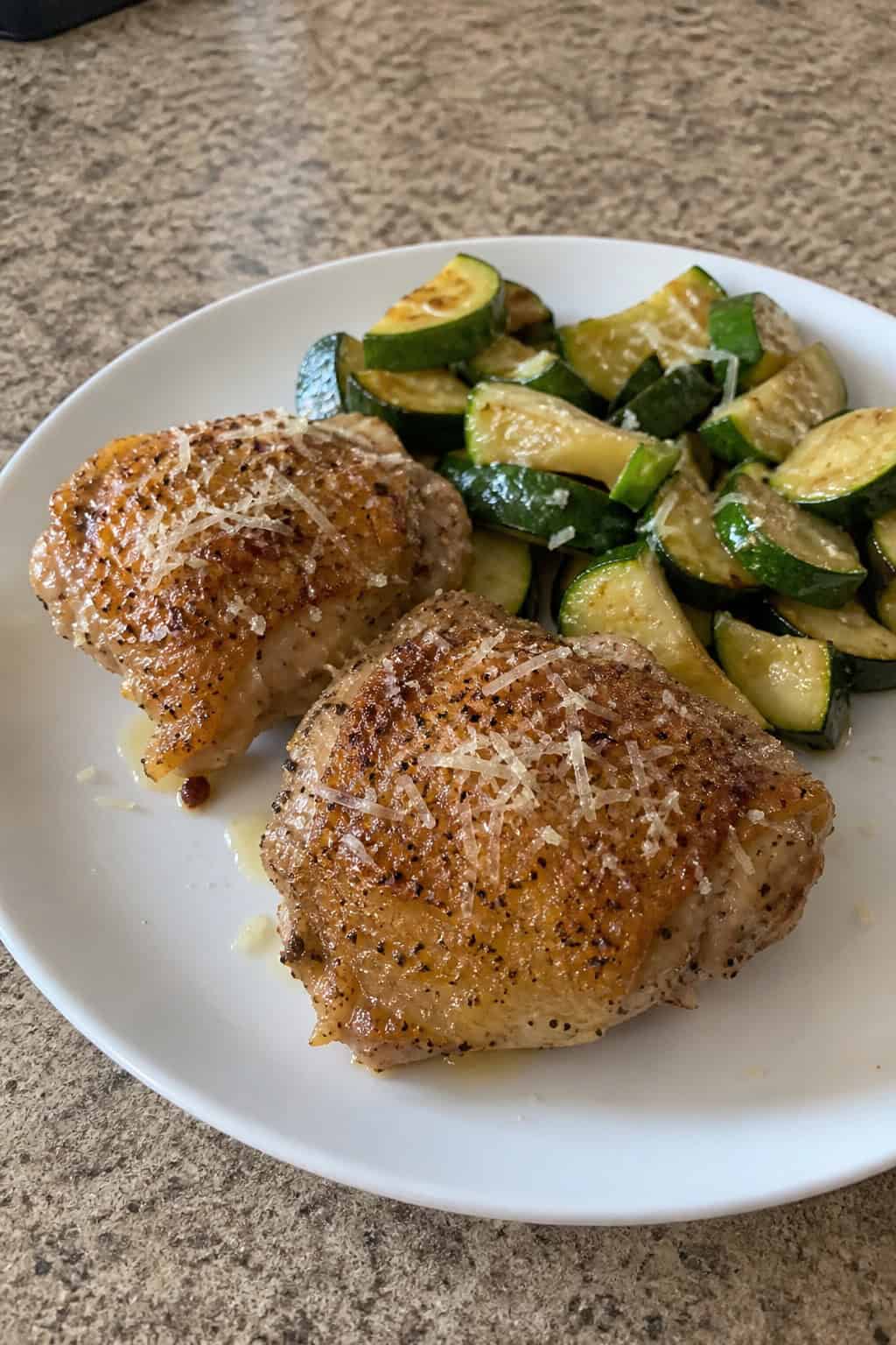 A plate with golden brown garlic parmesan chicken thighs served with sautéed zucchini on a wooden table.