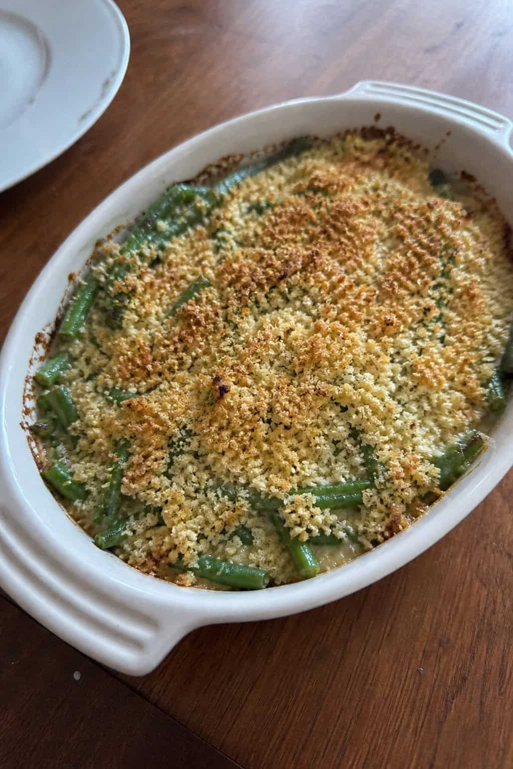 A garlic-parmesan green bean casserole with a golden panko crust in a white ceramic dish, with whole garlic cloves, a wedge of parmesan, and fresh herb sprigs arranged on the wooden table beside it.