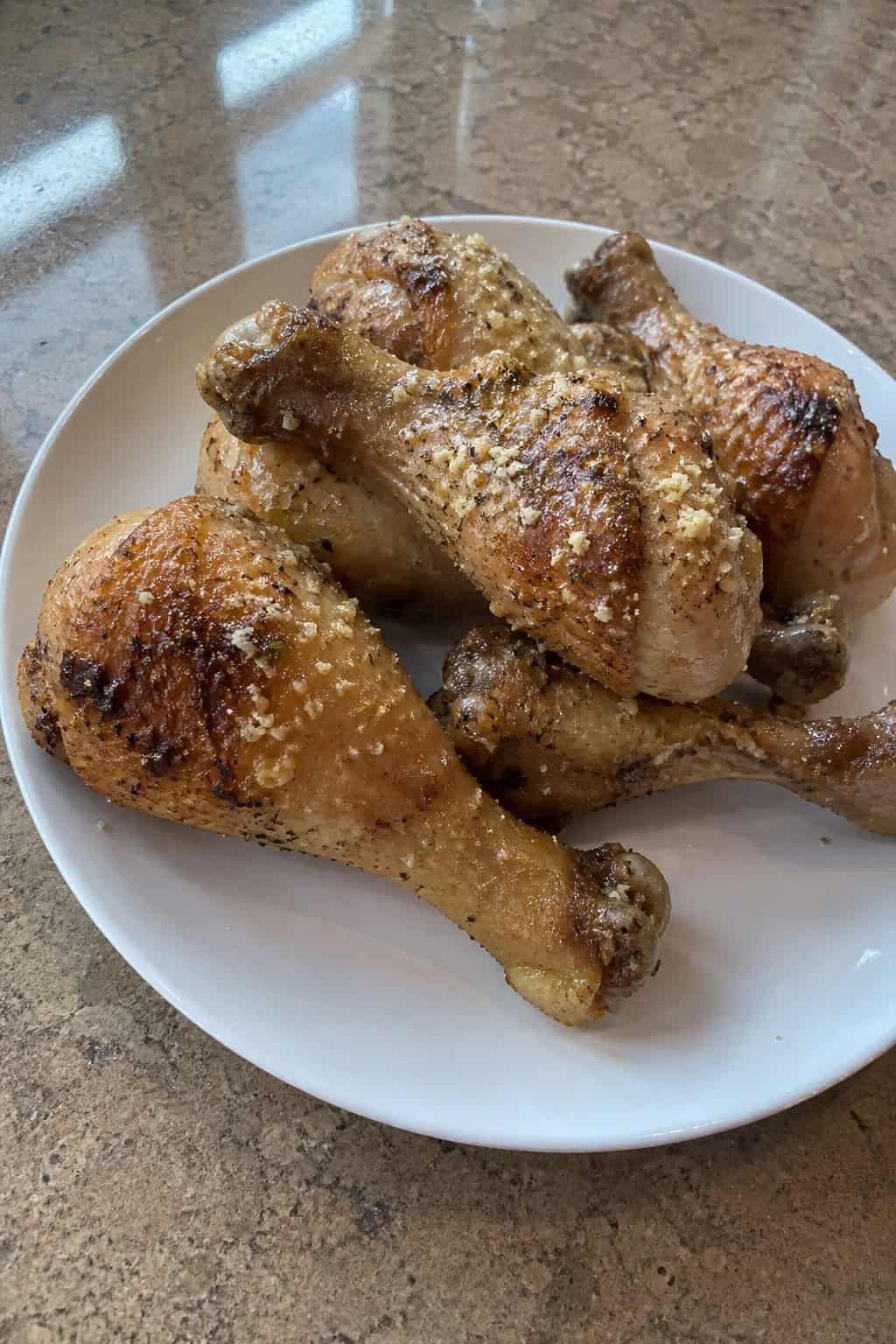 Golden garlic-Parmesan chicken drumsticks on a white plate topped with bright green parsley, with a small white bowl of creamy dipping sauce on a light kitchen table.