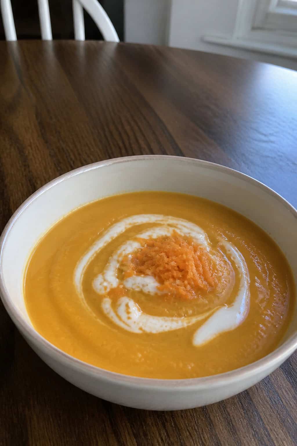 A bowl of ginger-carrot soup topped with yogurt and orange zest, surrounded by fresh carrots, ginger, and orange slices on a wooden table.