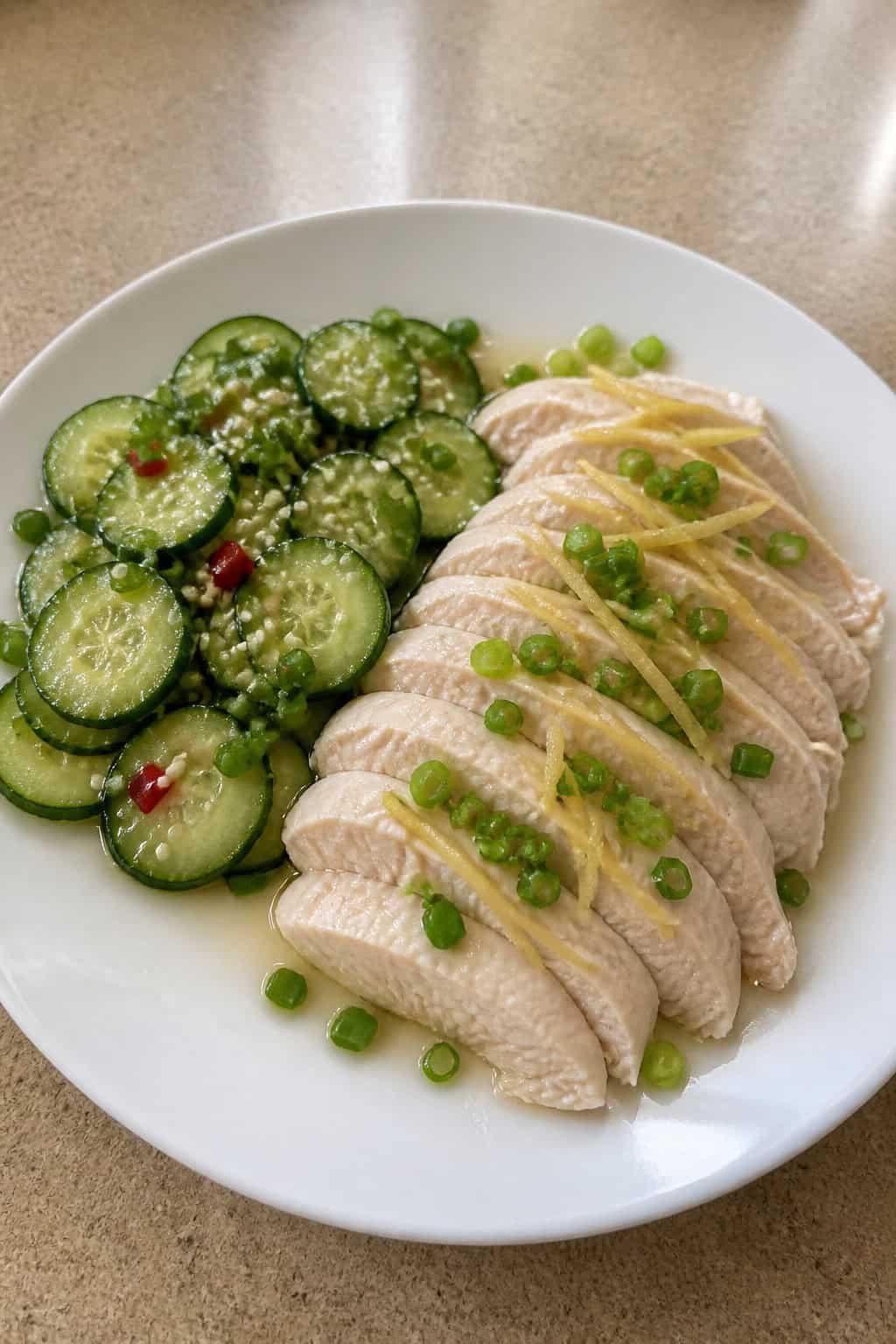 A plate of ginger-scallion poached chicken with sliced cucumber salad on a wooden table.
