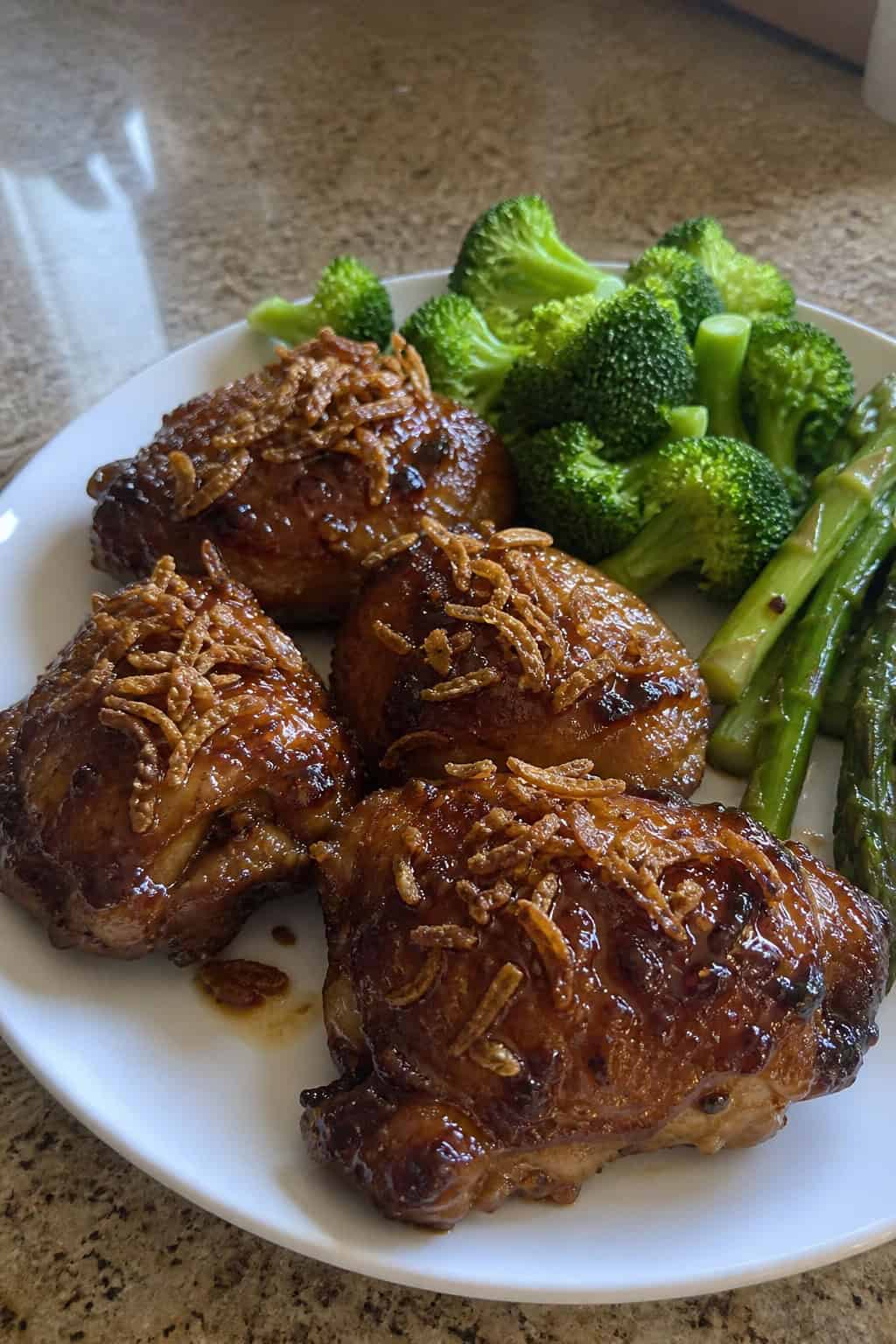 A plate of glazed honey garlic chicken thighs topped with crispy shallots and served with green vegetables.