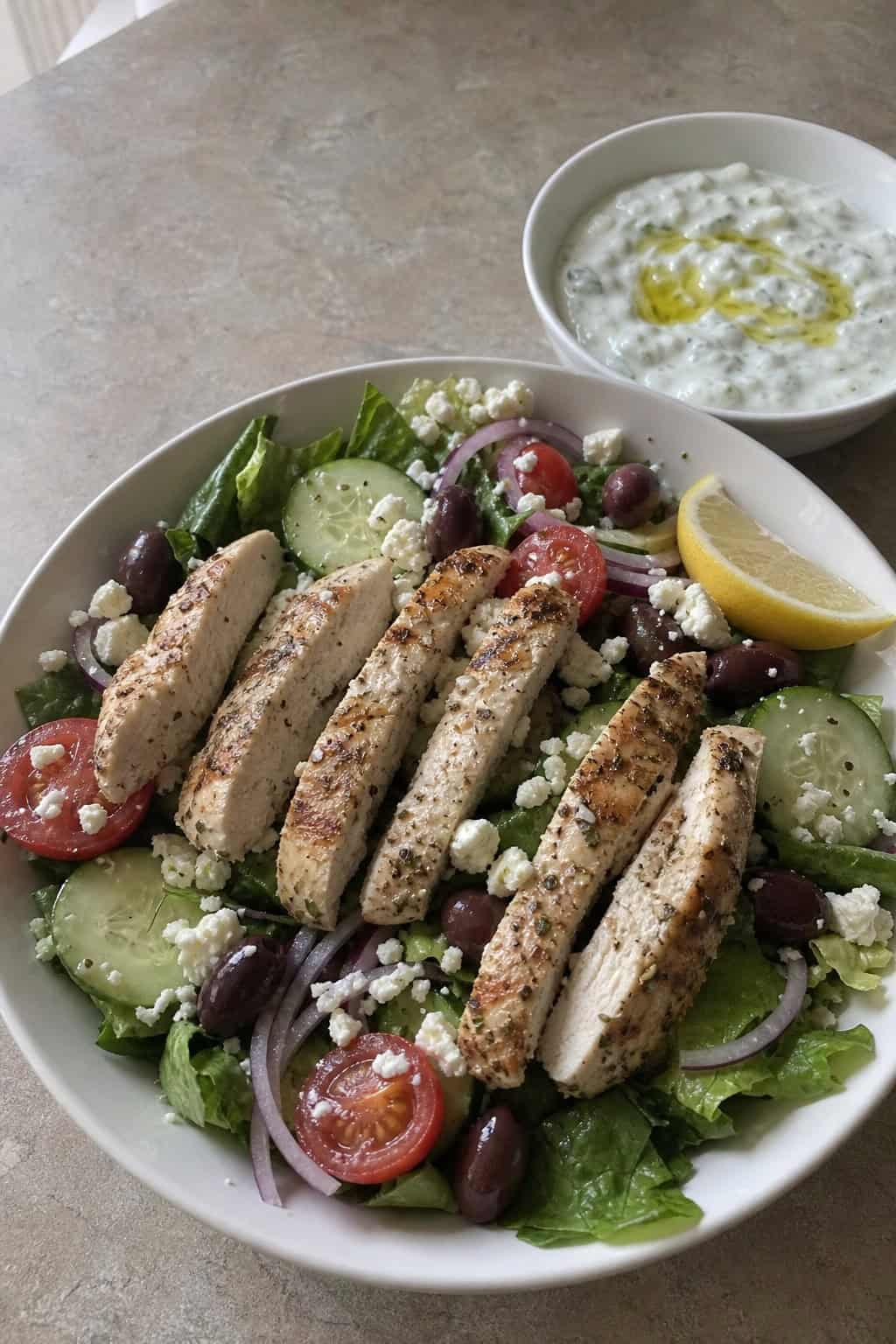 A large bowl of Greek salad with shredded chicken, chopped romaine, halved cherry tomatoes, diced cucumber, sliced red onion, crumbled feta, and Kalamata olives, with a small ramekin of tzatziki on the side.