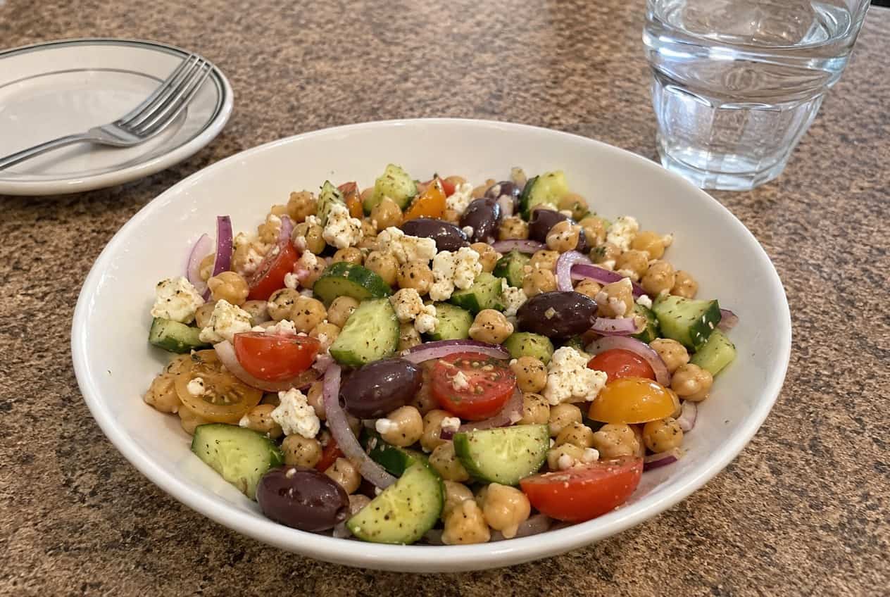 A large bowl of Greek chickpea salad packed with golden chickpeas, halved cherry tomatoes, diced cucumber, sliced Kalamata olives, crumbled feta, and scattered fresh parsley on a wooden table.