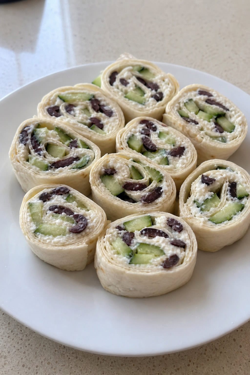 Plate of Greek feta, cucumber, and olive pinwheel appetizers on a wooden serving board showing white crumbled feta, thin cucumber rounds, and dark kalamata olives inside sliced flour tortilla rolls, with small bowls of olives and feta nearby.