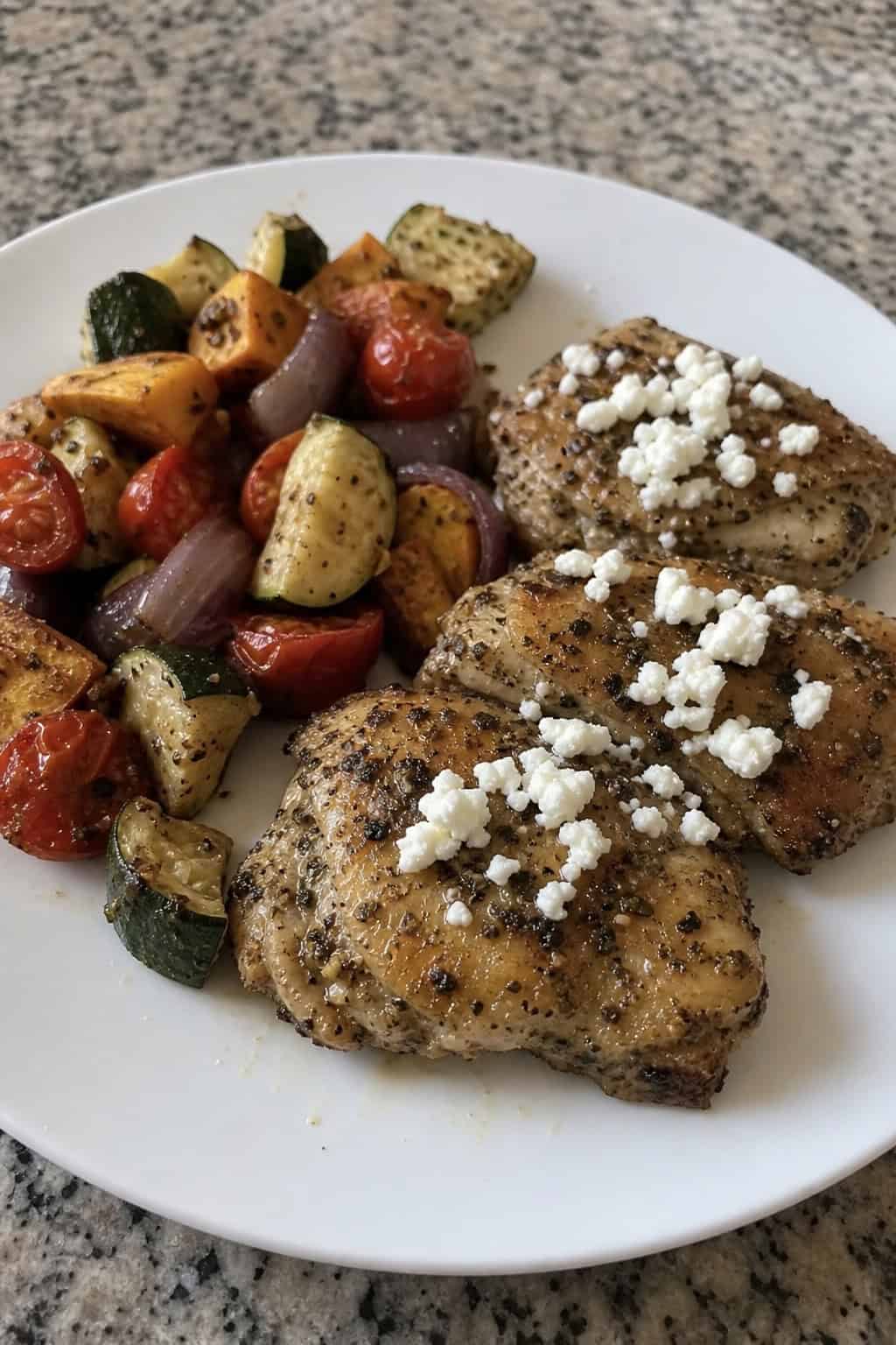 A plate of golden brown Greek lemon oregano chicken thighs topped with crumbled feta cheese and garnished with lemon wedges and fresh oregano, served with roasted vegetables.