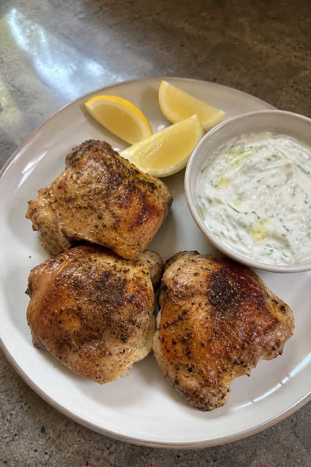 Sliced lemon-oregano chicken thighs on a wooden table with a small white bowl of thick tzatziki sauce, lemon wedges, and scattered fresh herb leaves nearby.